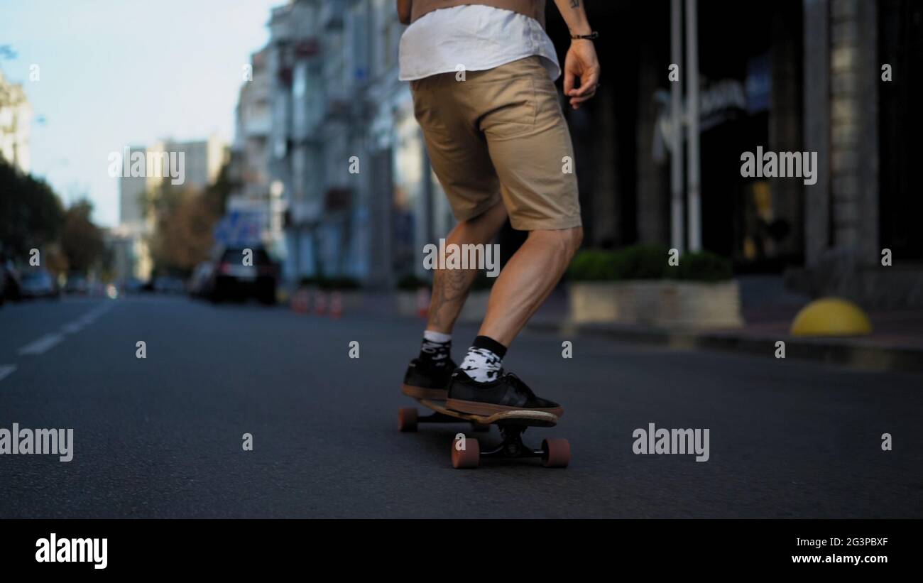 Un homme intelligent à la peau légère est passionné par la conduite rapide sur une longue planche sur la route. Les jambes sont rapprochées. Banque D'Images