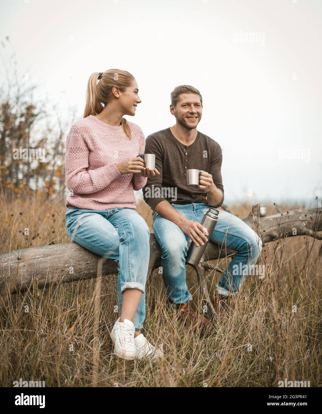 Un couple souriant de touristes boivent du thé et parlent tout en s'asseyant sur un arbre tombé au milieu d'une nature d'automne dehors Banque D'Images