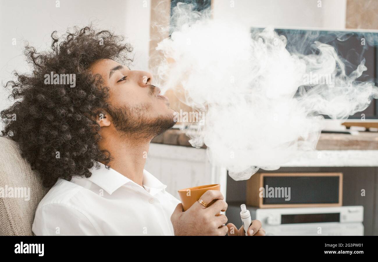 Jeune homme fume de la cigarette électronique à la maison. Le fumeur exhache un gros nuage de fumée, une vue prifile de l'arabe fume de boire Banque D'Images
