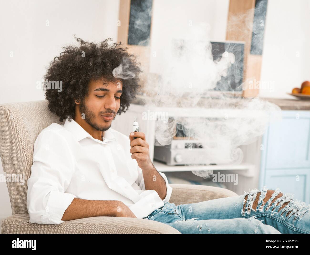 Homme barbu appréciant des bouffées de fumée de cigarette électronique. Un jeune homme dans une ambiance décontractée se repose à la maison assis dans un fauteuil et dans un fumoir Banque D'Images