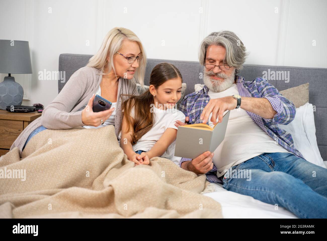 Petite petite petite fille petite-fille souriant lors de la lecture d'un livre avec des grands-parents attentionnés tout en étant allongé sur un lit spacieux et en appréciant le temps libre avec grand-mère et grand-père le week-end à la maison. Concept de famille Banque D'Images