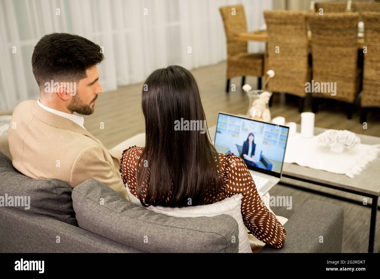Une jeune femme et un mari parlent de problèmes avec un psychologue de famille par appel vidéo sur un ordinateur portable tout en étant assis sur un canapé à la maison, vue arrière. Jeune couple ayant consultation en ligne avec le médecin Banque D'Images