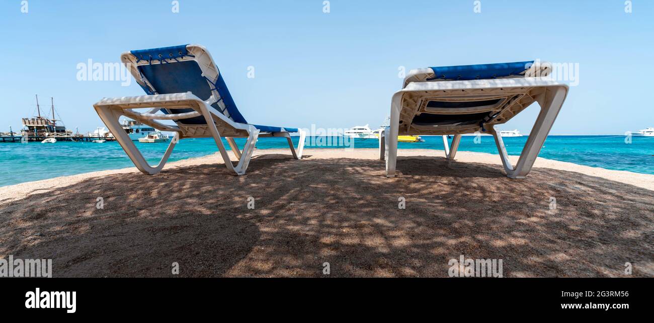 Deux chaises longues de plage à l'ombre sur les rives de la mer Rouge avec la toile de fond des bateaux et des navires en Egypte Hurghada Banque D'Images