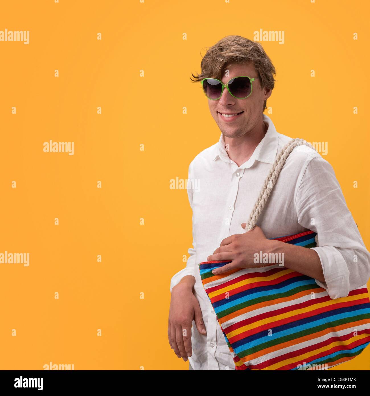 Jeune homme drôle avec grand sac multicolore se réjouit regardant à travers des lunettes de soleil. Découper sur fond jaune. Concept commercial Banque D'Images