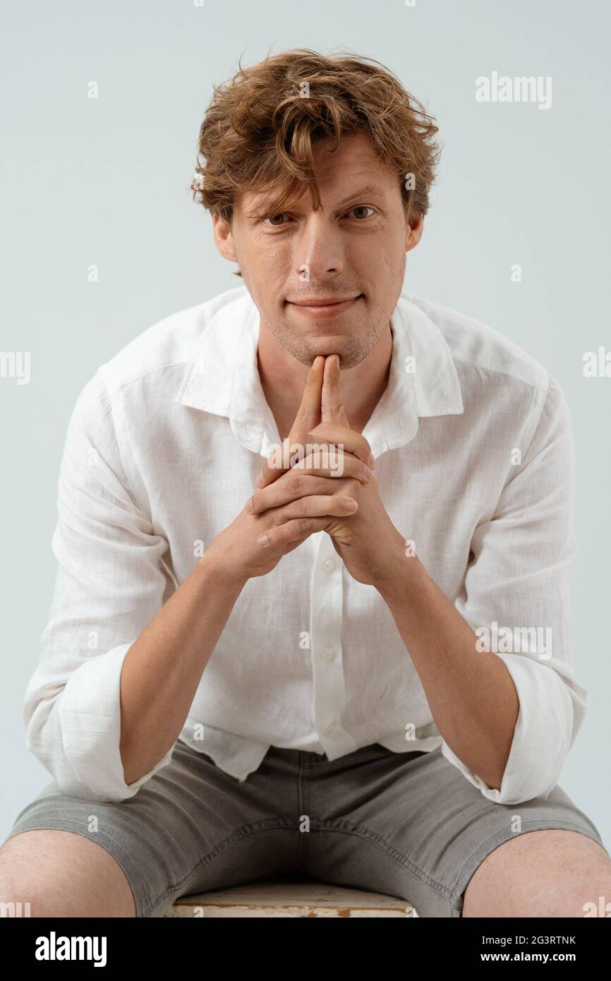 Sourires beaux touchant avec les index de son menton. Portrait d'un adorable gars à cheveux rouges en chemise blanche Banque D'Images