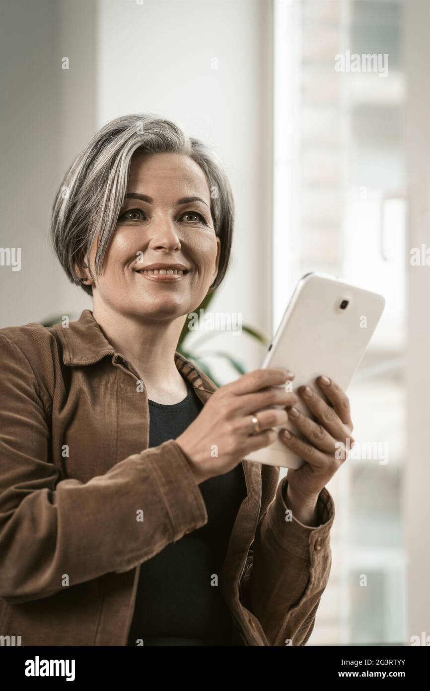 Une jolie femme en âge de prendre sa retraite sourit de communiquer en ligne sur une tablette numérique tout en se tenant près de la fenêtre. Concept de technologie Banque D'Images