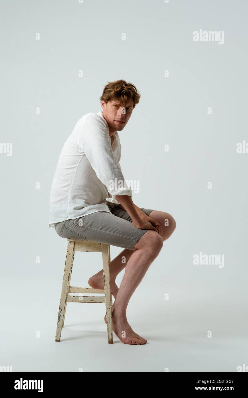 Homme pieds nus assis sur une chaise. Vue latérale d'un jeune homme en chemise blanche et Jean sur fond blanc. Banque D'Images