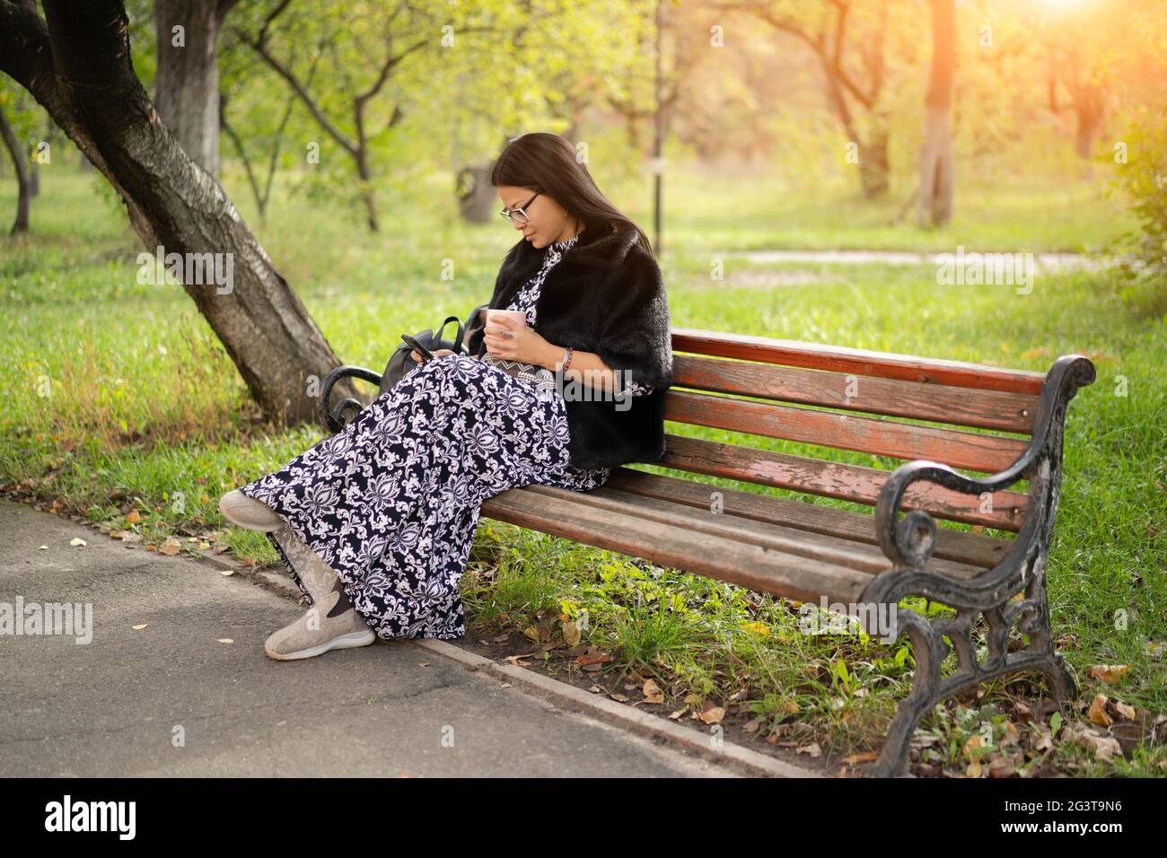 Femme d'origine asiatique dans une belle longue robe s'assoit au parc sur un vieux banc rouillé et lit le livre en utilisant son gadget ou chèques socia Banque D'Images