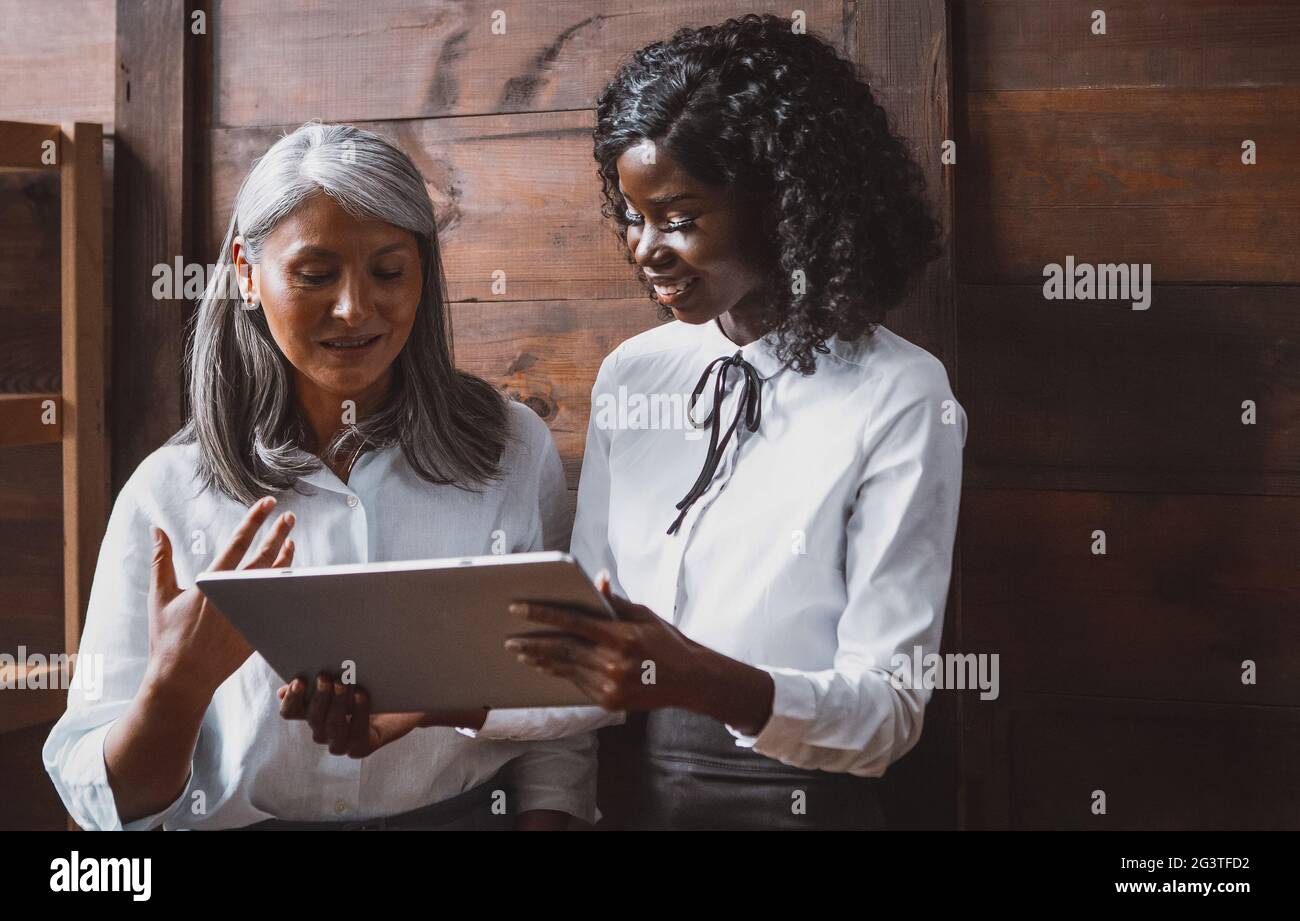 Femme asiatique mûre et jeunes femmes d'affaires africaines travaillant ensemble à côté d'un mur de bureau en bois tenant un onglet numérique Banque D'Images