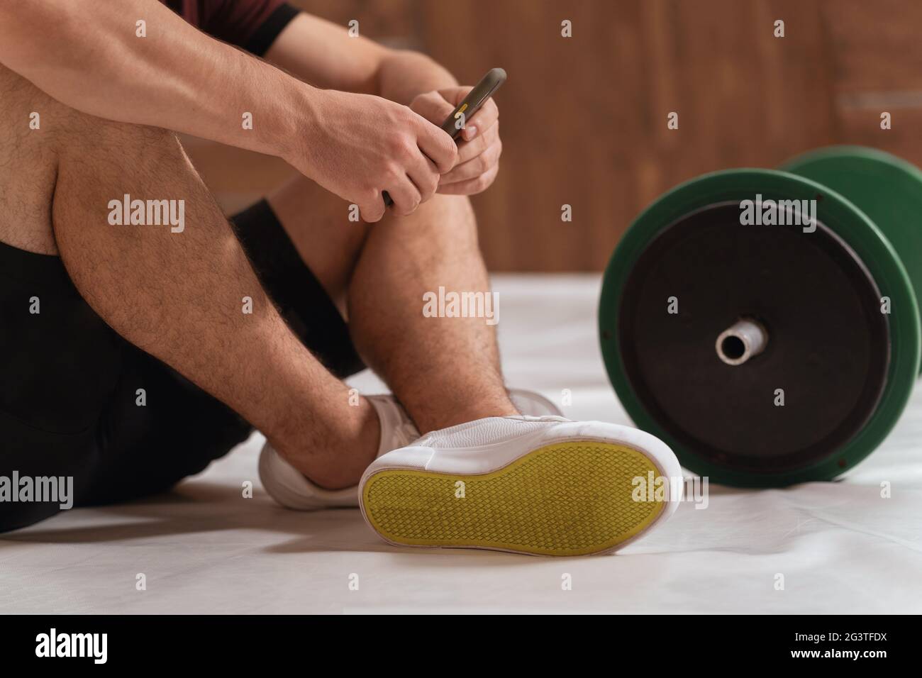 Gros plan. Jeune homme ayant une pause assis sur un sol avec une barbell de fitness noir et vert, équipement pour l'entraînement de poids c Banque D'Images