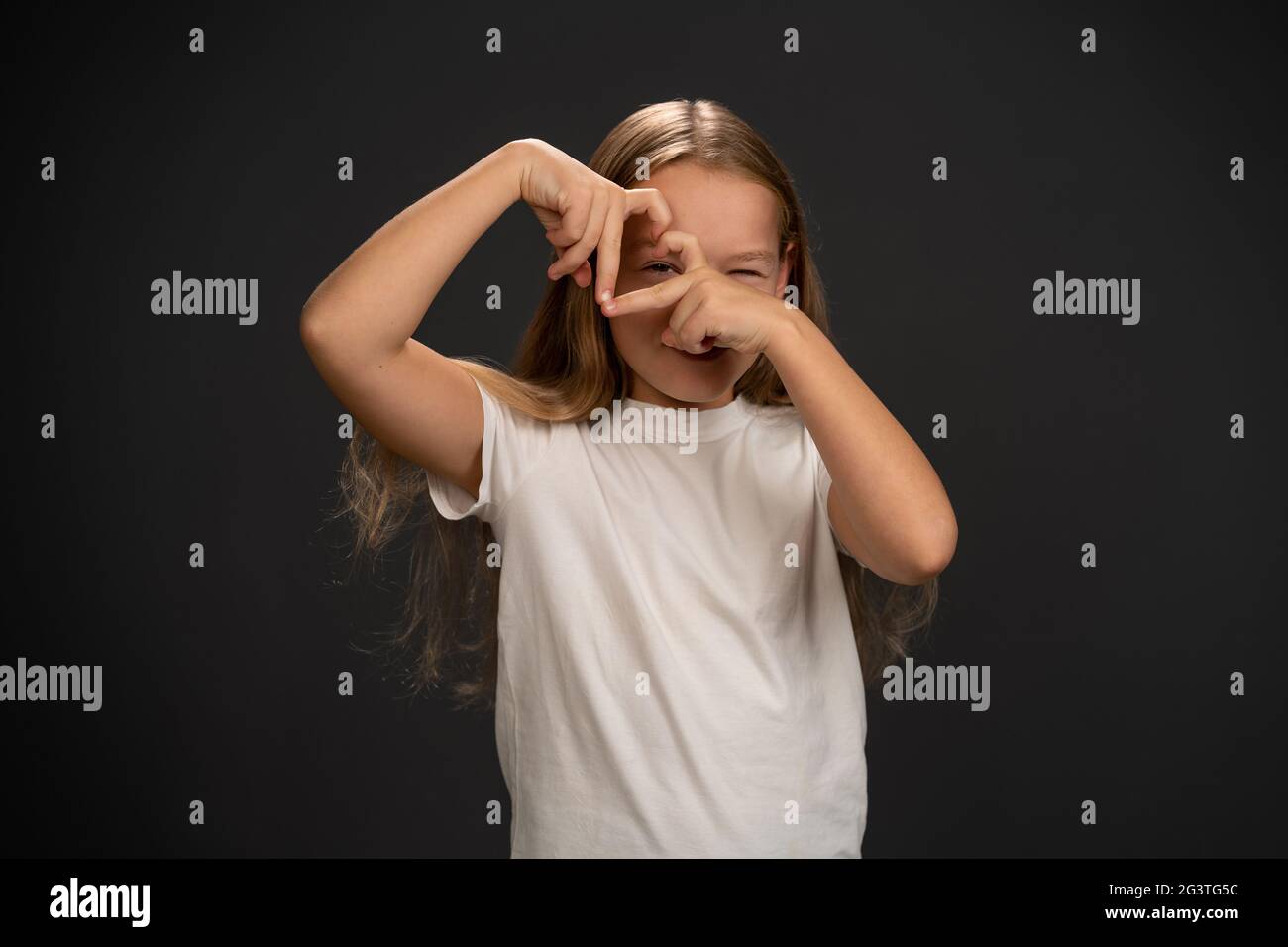 Amour geste heureuse fille de 8,10 ans tenant ses mains ensemble faisant une forme de coeur et regardant à travers elle portant t-shirt blanc Banque D'Images