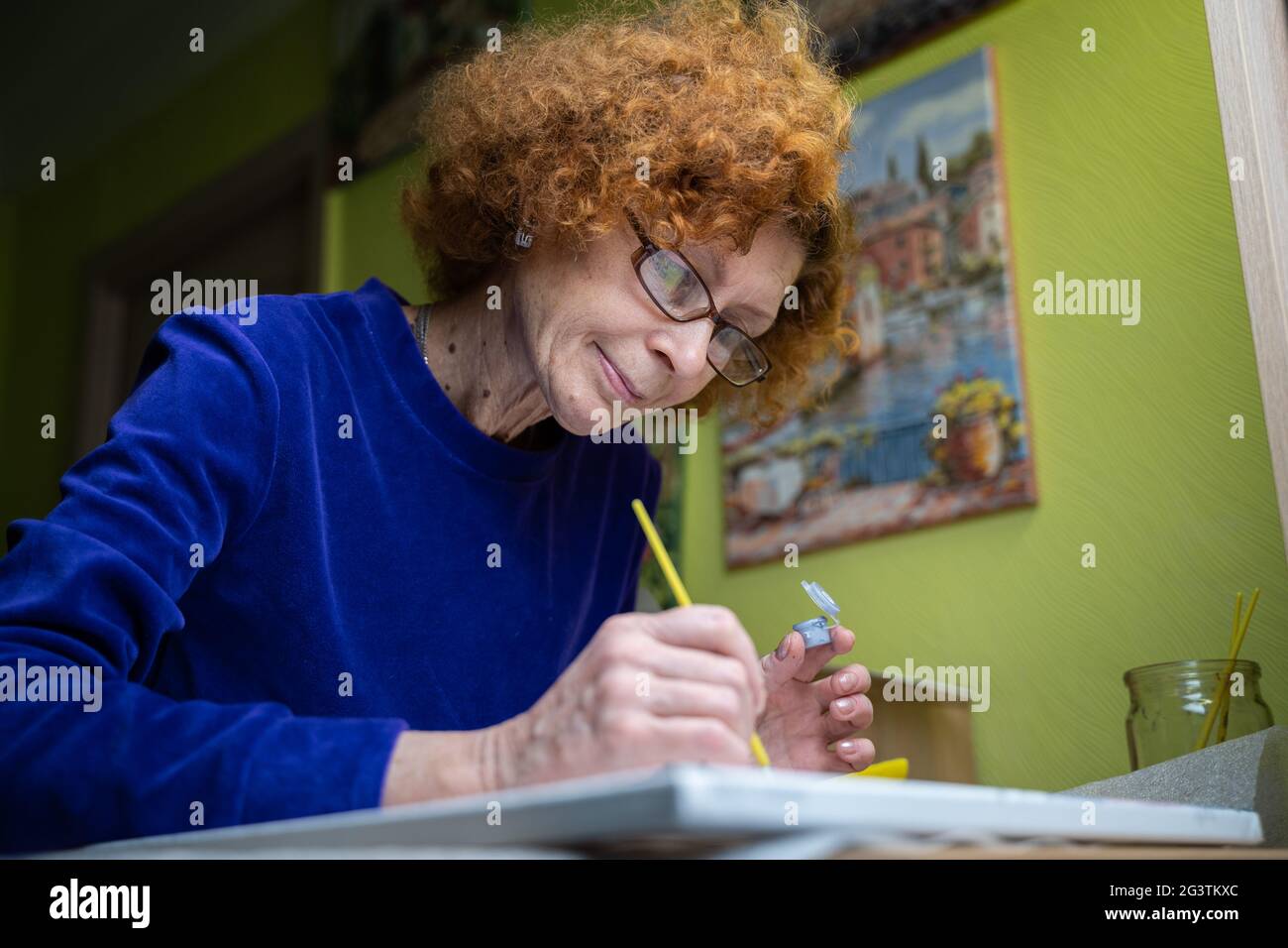 Bonne femme d'artiste mûre peint sur toile à la maison studio. Exprimez votre créativité. Femme âgée positive tenant la brosse et le tirage Banque D'Images