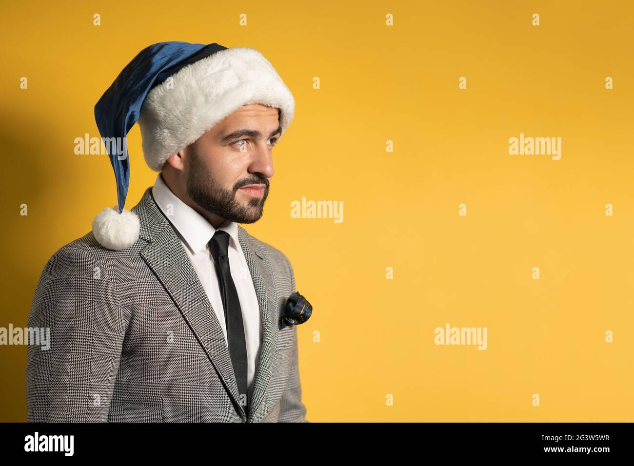 Charmant jeune homme barbu en chapeau bleu de Santa et costume gris, en regardant latéralement isolé sur fond jaune. Joli émo pensif Banque D'Images