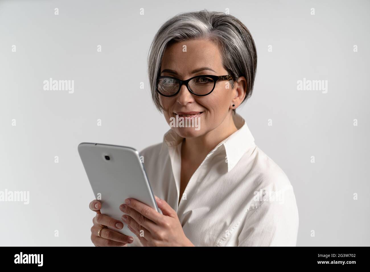 Portrait d'une femme mûre aux cheveux gris en chemise blanche et lunettes à l'aide d'une tablette numérique. L'utilisation de la technologie par les personnes âgées Banque D'Images