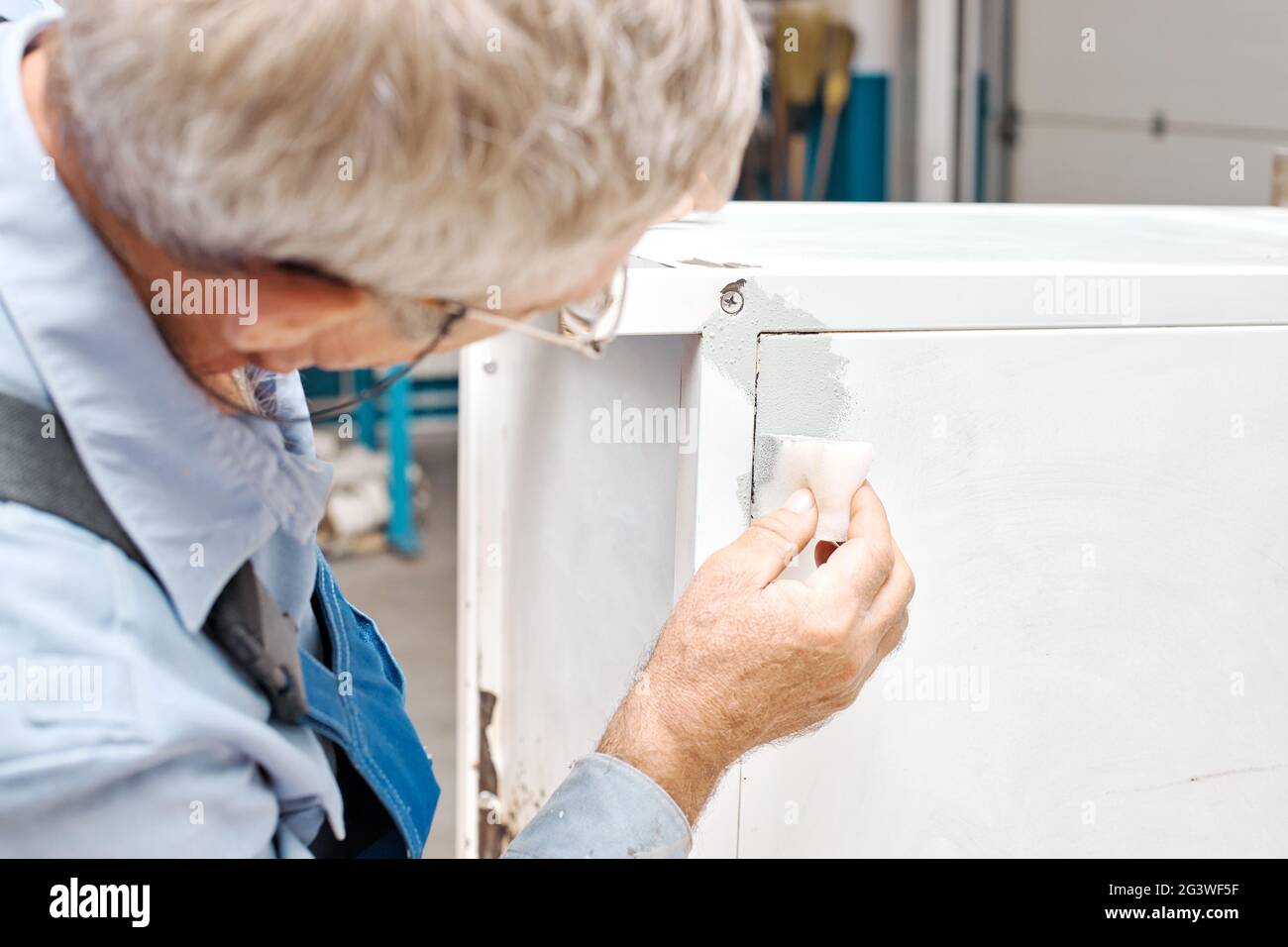 Un employé peint des produits métalliques. Un homme âgé tient une éponge en mousse dans sa main et peint le cabinet en blanc. Banque D'Images
