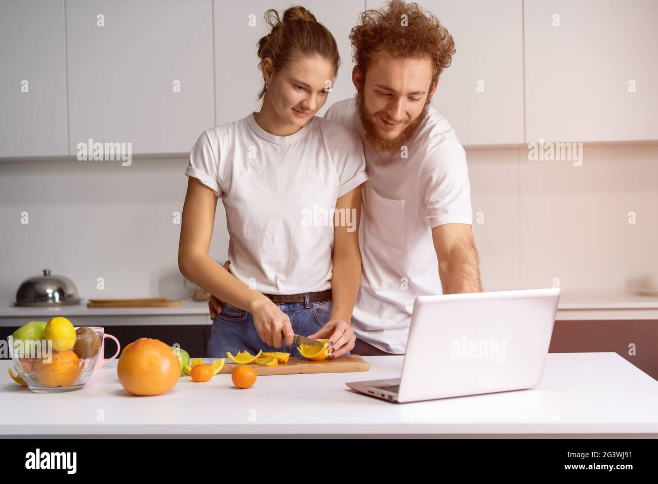 L'homme se pencha sur une fille souriante regardant un film romantique. Beau jeune couple parlant sur appel vidéo à l'aide d'un ordinateur portable. Jeune couple cuisinier Banque D'Images