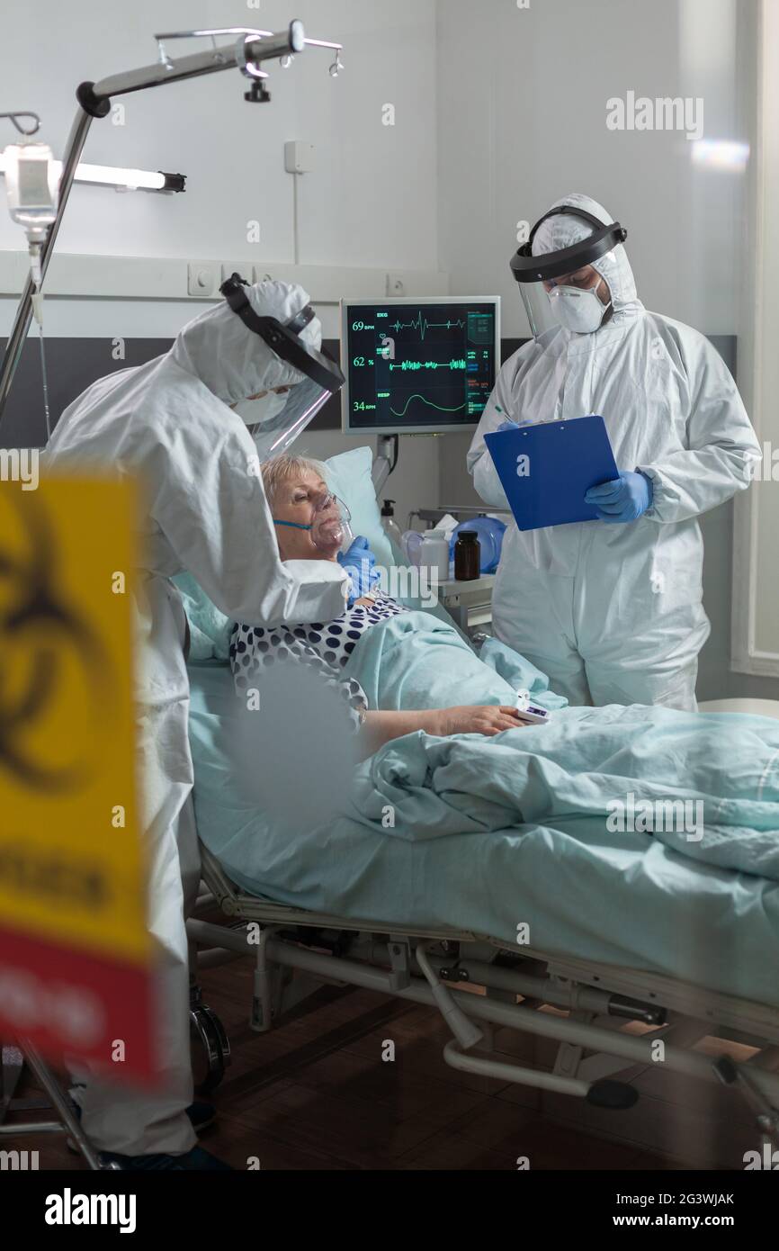 Une infirmière médicale vêtue d'un équipement de protection individuelle qui met un masque à oxygène au patient âgé, au cours d'une pandémie mondiale avec coronavirus, et le médecin prend des notes sur le presse-papiers. Banque D'Images