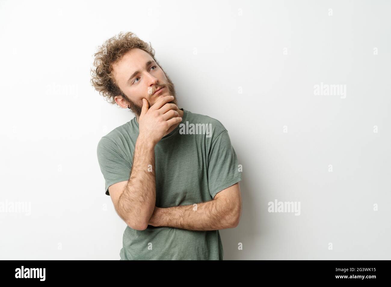 Regard pensif d'un jeune homme beau avec des cheveux bouclés dans un t-shirt olive, en regardant vers le haut, isolé sur fond blanc. Portrait de Banque D'Images