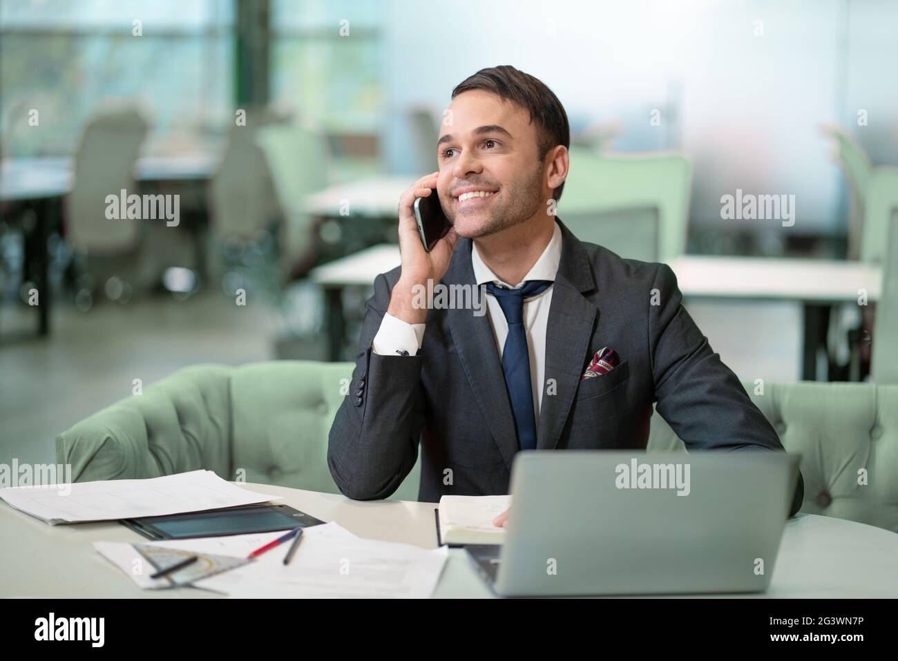 Beau homme en costume de travail parlant sur le téléphone de travail sur ordinateur portable, travail en cours. Encouragez le freelancer souriant à prendre des notes Banque D'Images