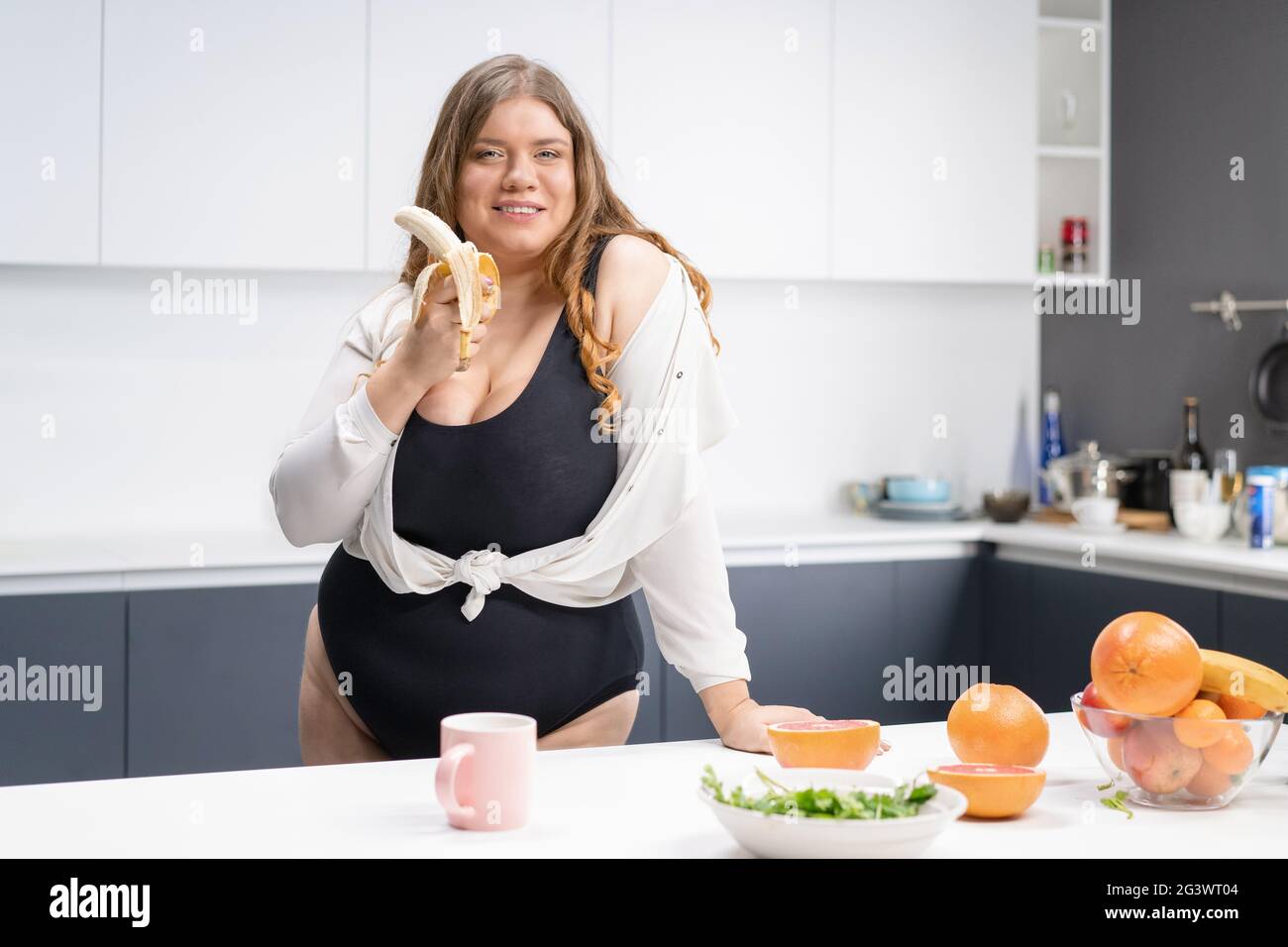 Concept de régime et de nutrition. Joyeux curvy corps jeune femme avec de longs cheveux blonds en utilisant le blender dans la cuisine moderne, en mélangeant frais Banque D'Images