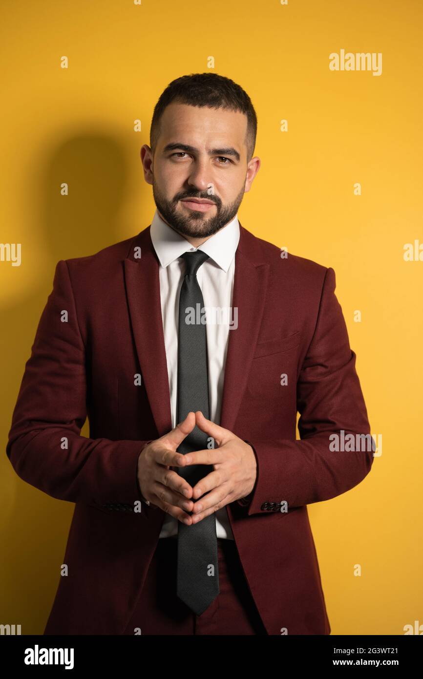 Homme d'affaires prospère. Portrait d'un beau homme en couleur bordeaux costume regardant l'appareil photo avec les mains repliées isolé sur jaune b. Banque D'Images
