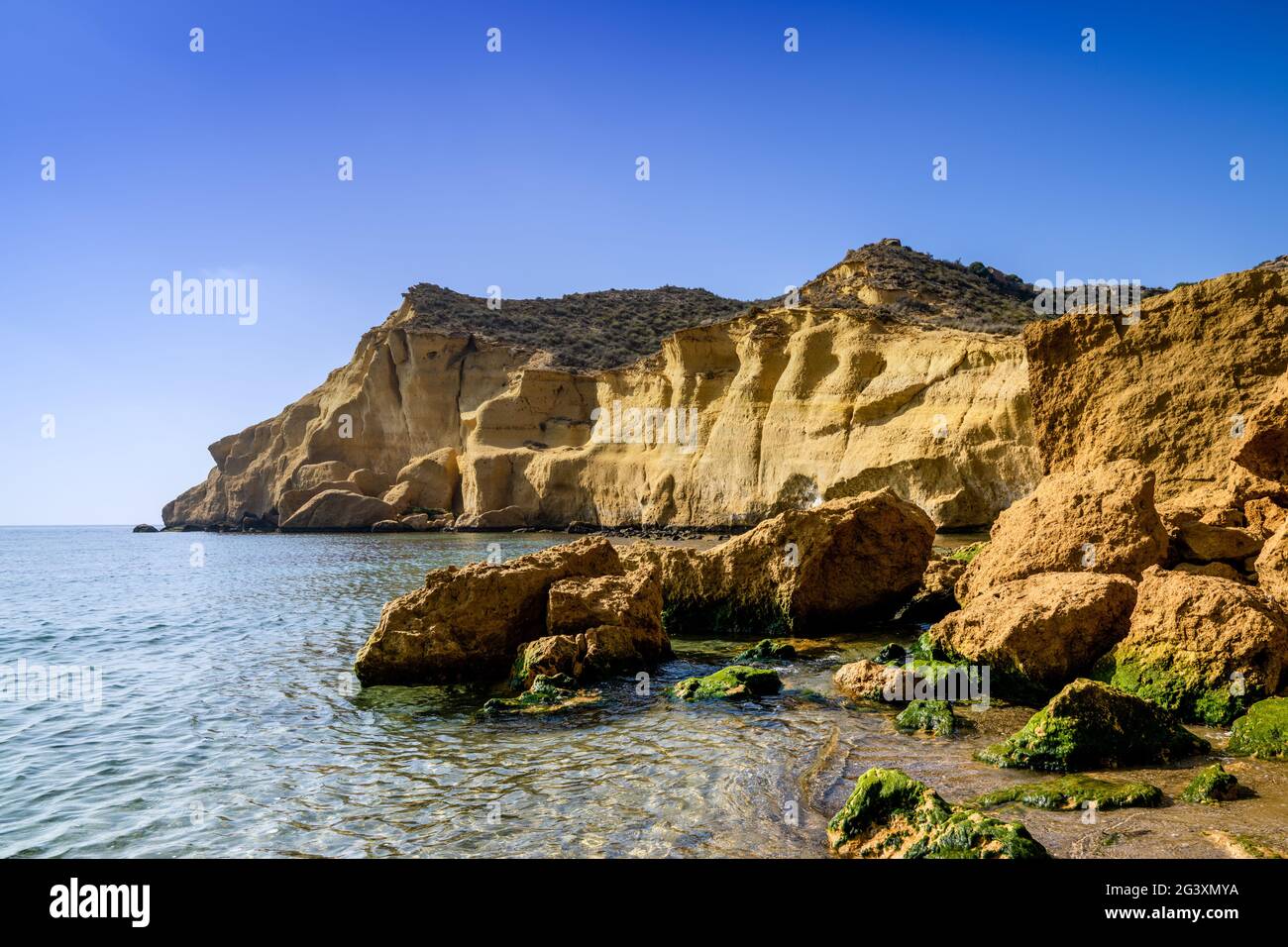 Une crique paisible et idyllique de l'océan dans la Méditerranée avec des falaises de grès jaune derrière et des rochers couverts d'algues vertes sur le sable Banque D'Images