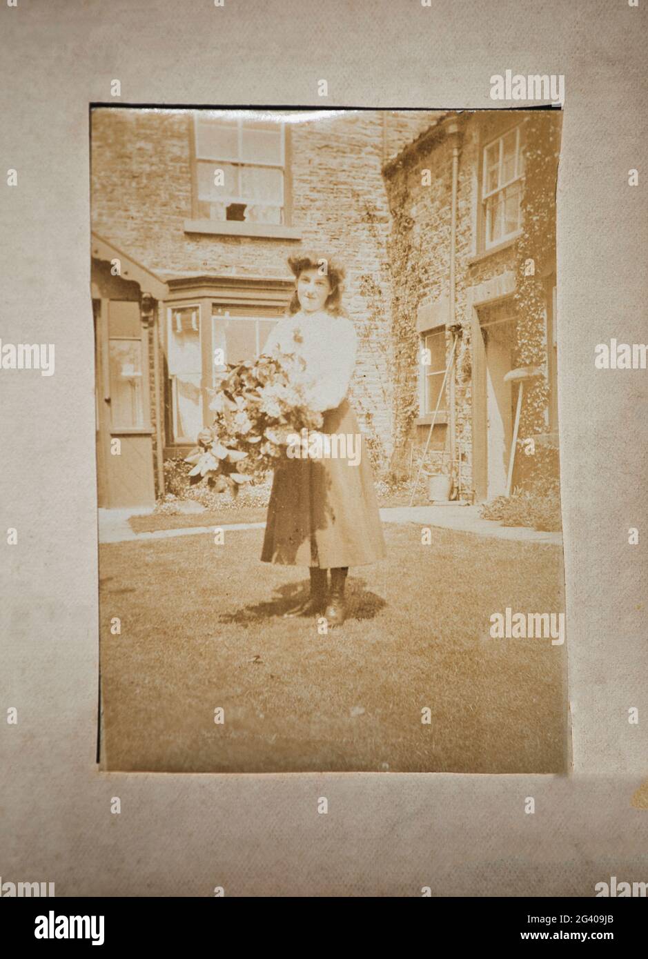 Photographie sépia authentique de jeune femme souriante portant des fleurs debout devant la maison. Concept de nostalgie, style de vie, historique Banque D'Images
