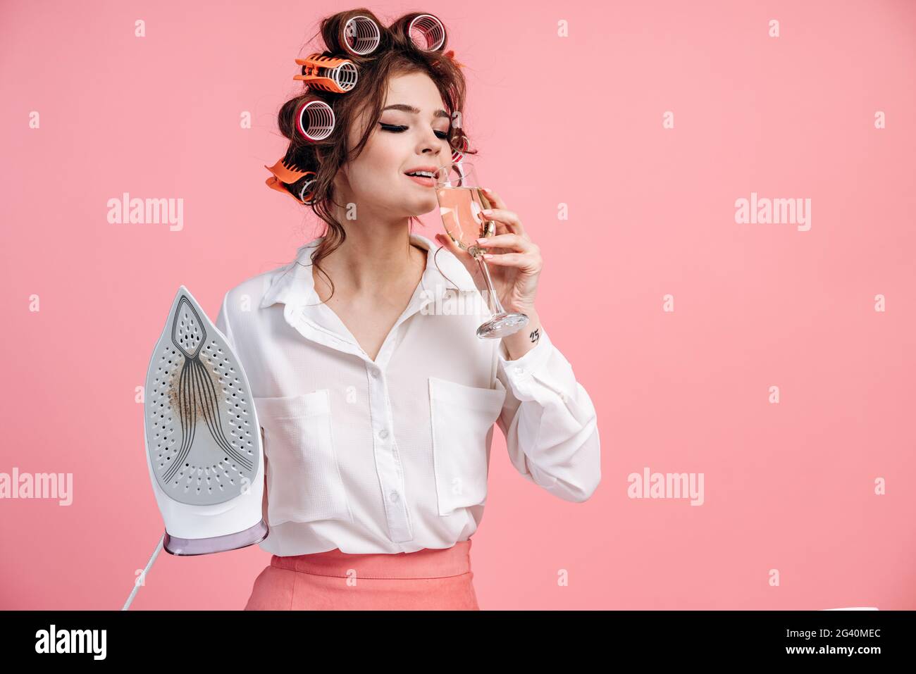 Élégante brune, femme au foyer avec un verre d'alcool et un fer sur fond rose. Belle fille bien entretenue avec des boucles sur ses cheveux. Banque D'Images