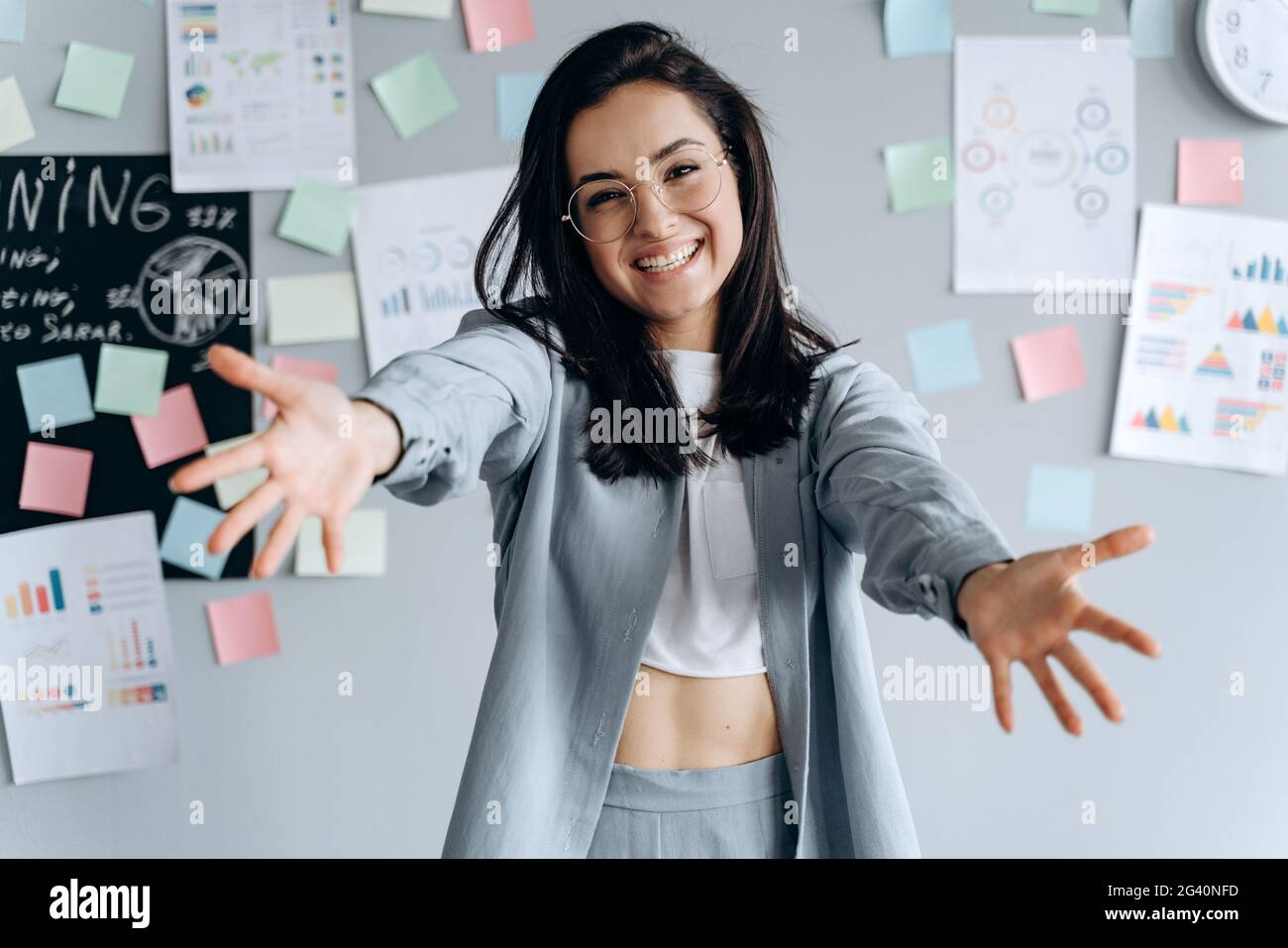 Belle jeune femme debout sur les murs gris, regardant l'appareil photo, souriant avec les bras ouverts pour les câlins. Fille gaie dans le bureau. Banque D'Images