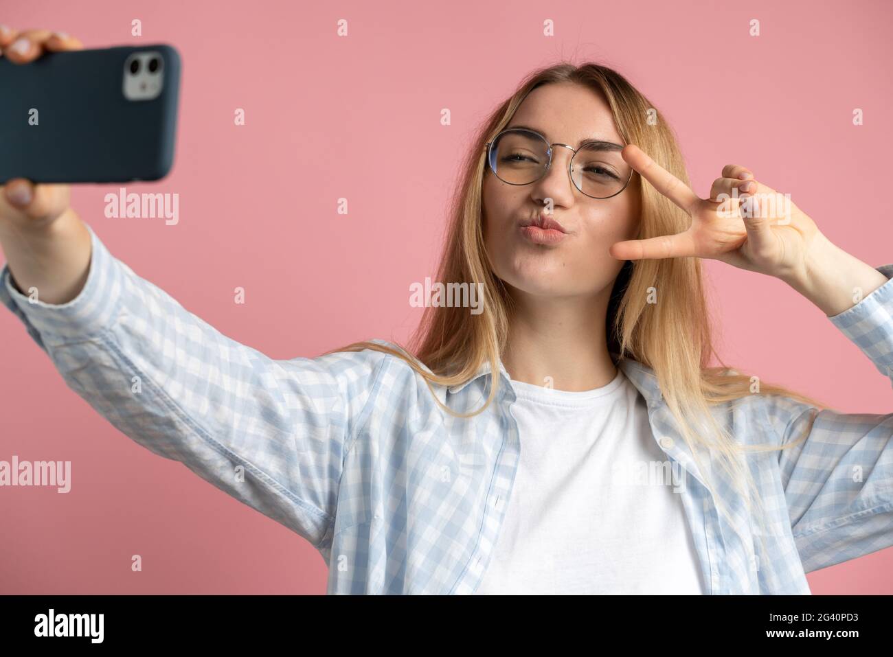 Positive fille avec des verres prend un selfie sur un fond rose. La jeune fille fait un geste de paix autour des yeux et un baiser. Banque D'Images