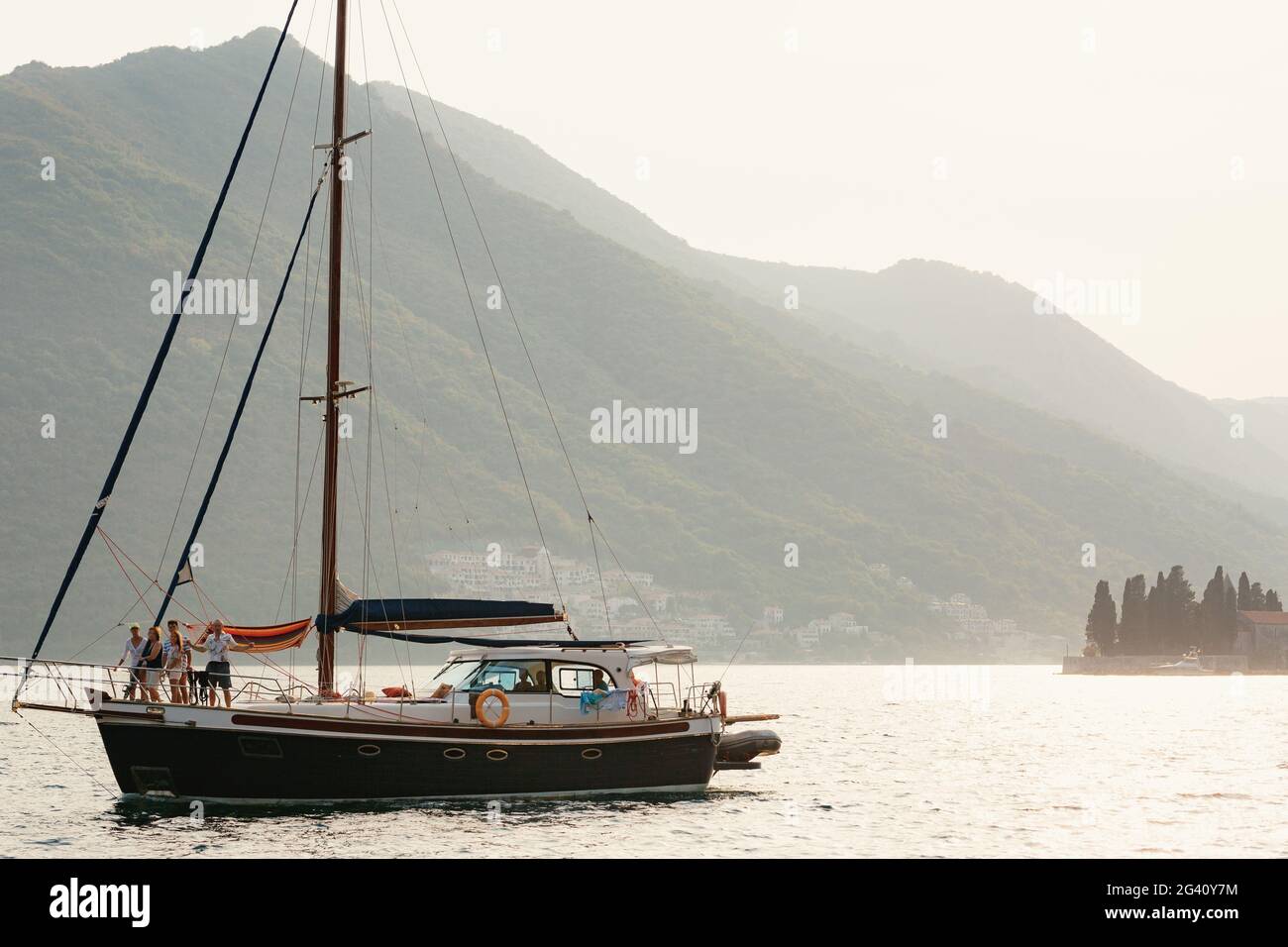 Un yacht avec des voiles abaissées et des personnes à bord avec la toile de fond des montagnes de la ville de Perast au Monténégro. Banque D'Images