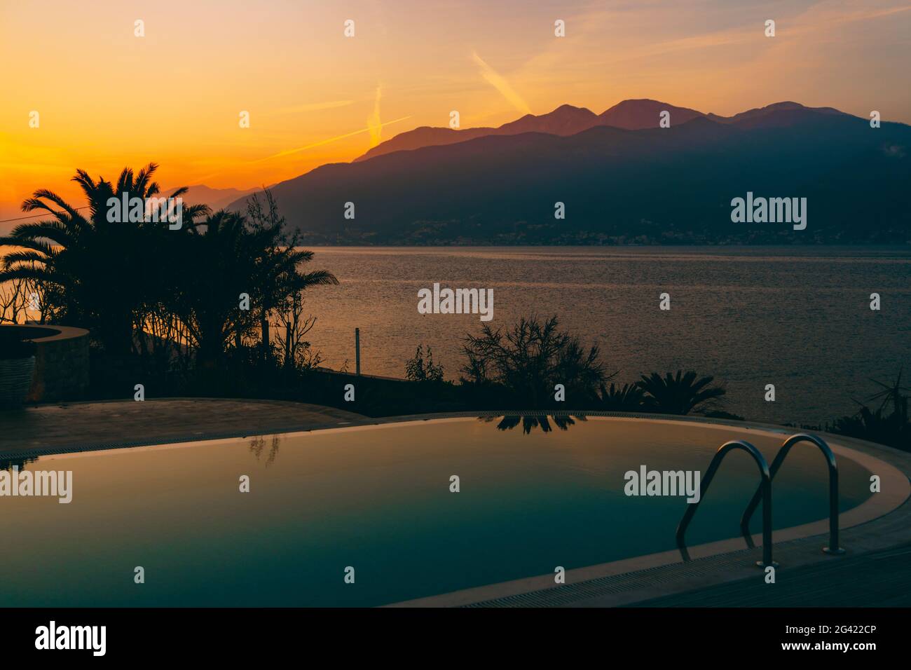 Le coucher de soleil se reflète dans la piscine, sur fond de mer, de montagnes et de palmiers. Banque D'Images