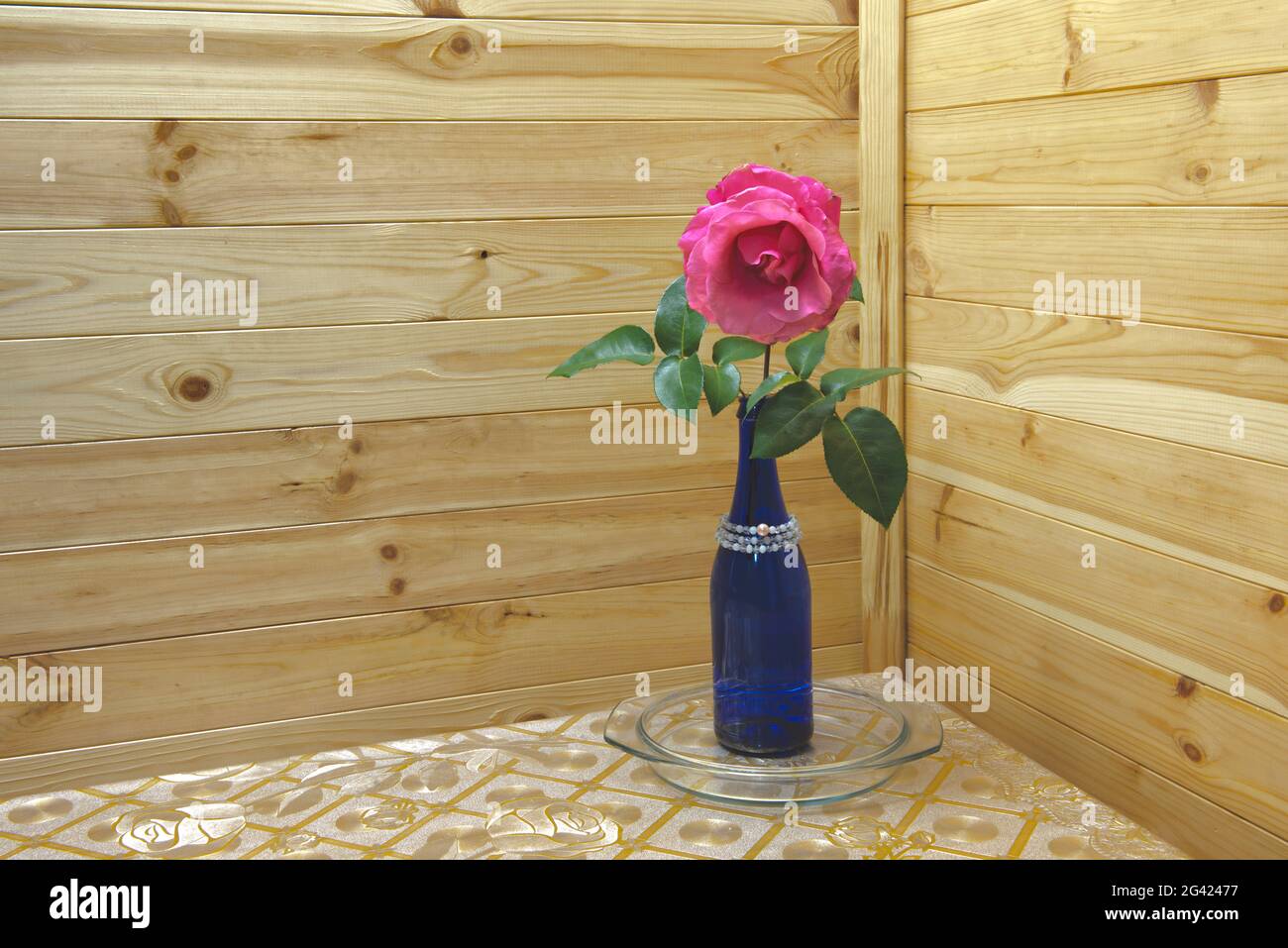 Une rose dans la bouteille bleue sur la table de cuisine dans le coin intérieur sur fond de mur de doublure en bois. Banque D'Images