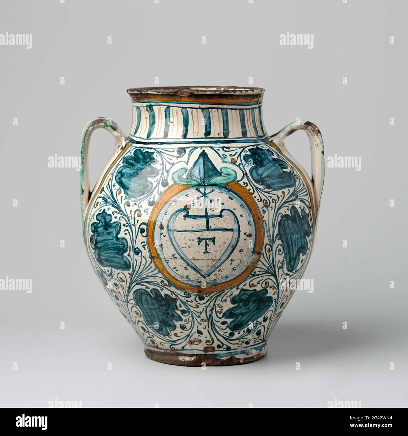 Vase, sphérique, avec deux oreilles. Vase sphérique de majolique peint multicolore, avec un col cylindrique court et deux oreilles en forme de C. Une arme en forme de coeur a été peinte dans un médaillon circulaire à l'avant du vase, à l'intérieur duquel la lettre t et au-dessus d'une croix. Autour du médaillon et à l'arrière du vase se trouvent des branches avec des spirales peintes entre elles de grandes fleurs stylisées bleues. Banque D'Images