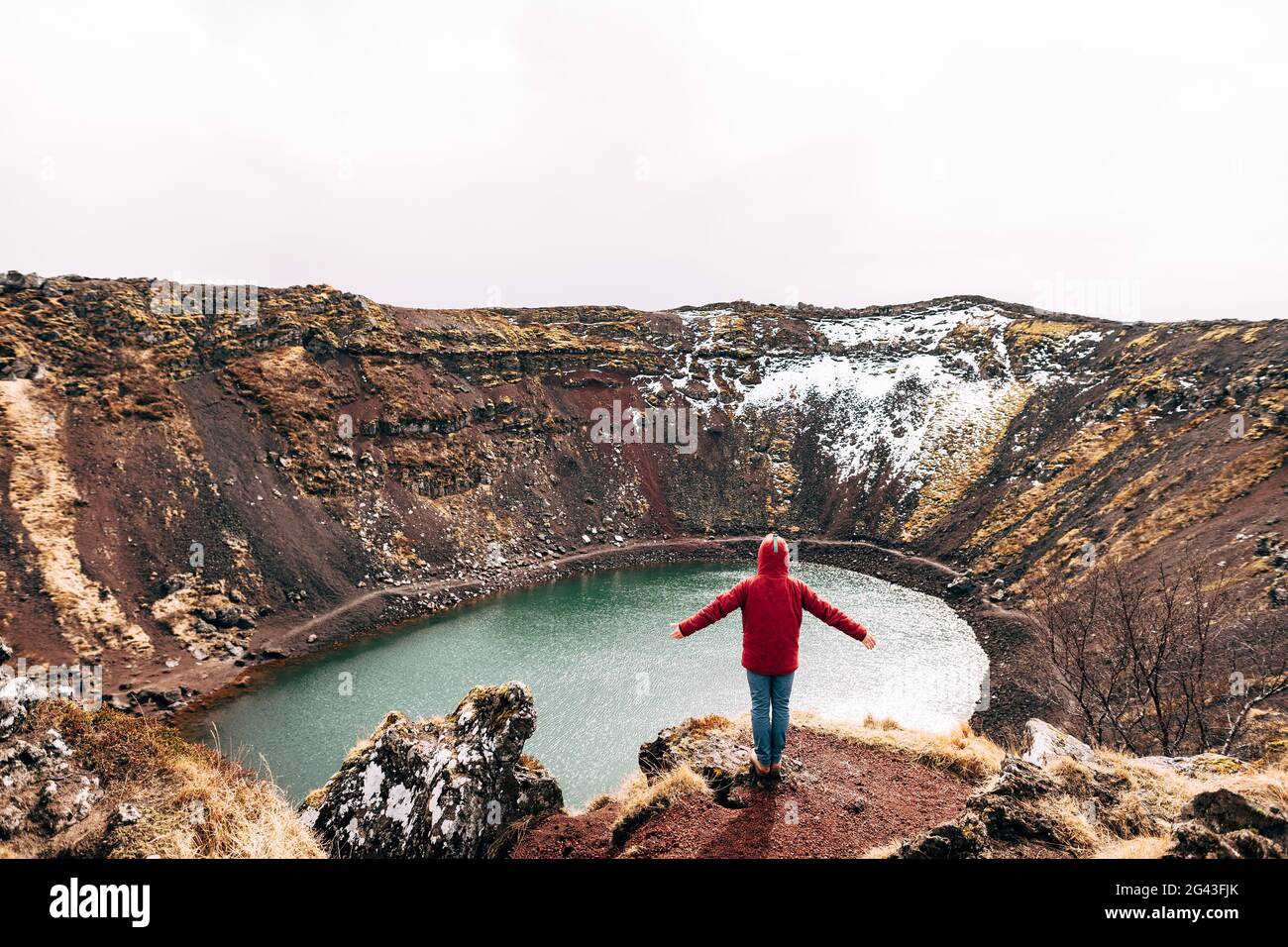 Un gars dans une veste rouge se dresse sur un rocher au-dessus du lac Kerid - un lac volcanique cratère en Islande. Sol volcanique rouge, similaire à Marti Banque D'Images