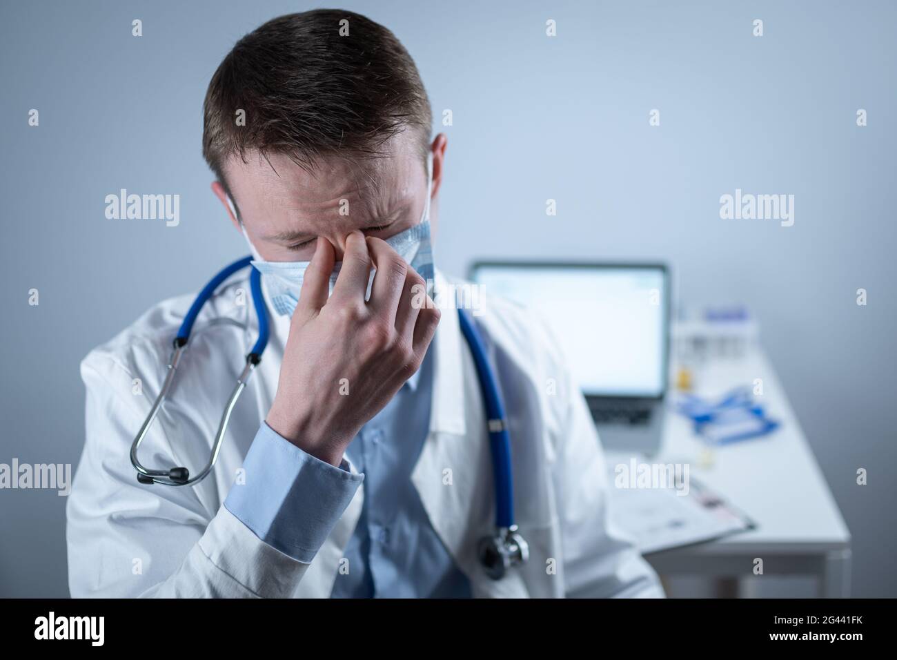 Jeune médecin européen allemand regardant l'appareil photo portant des lunettes et un masque dans le bureau de la clinique sur le lieu de travail avec le regard fatigué. Le sujet est Banque D'Images