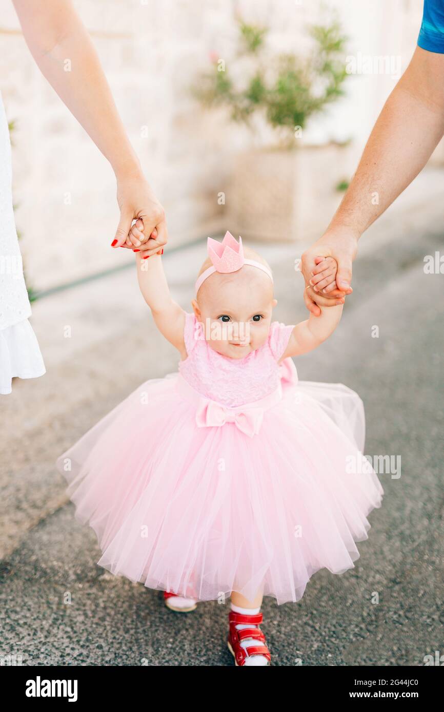 Une charmante petite fille dans une robe rose, des baskets et avec une couronne sur sa tête tient maman et papa à la main Banque D'Images