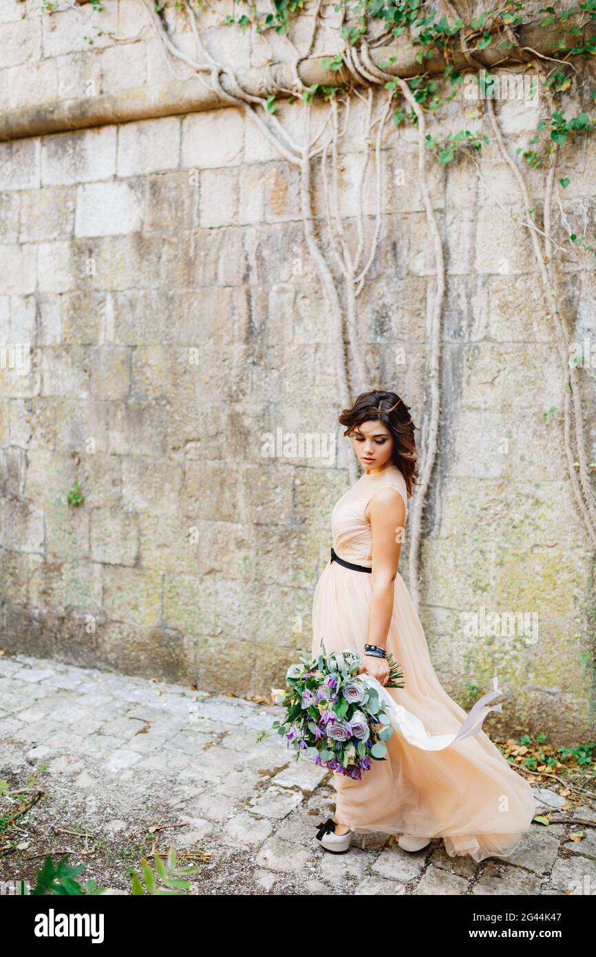 Belle mariée dans une robe pastel et de hauts talons chaussures marche avec un luxueux bouquet de fleurs le long d'un mur en pierre Banque D'Images