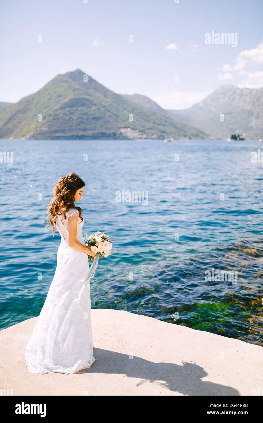 Une mariée élégante avec un bouquet dans ses mains se tient Sur un quai dans la baie de Kotor près de la eau bleue transparente Banque D'Images