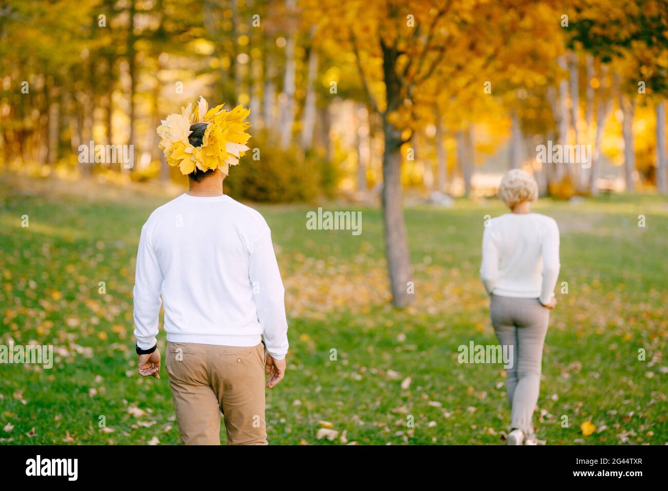 L'homme dans une couronne de feuilles jaunes suit la femme dans des vêtements légers dans la forêt d'automne. Vue arrière Banque D'Images