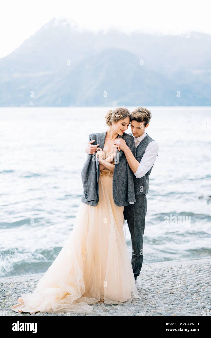 Marié jeta une veste sur les épaules de la mariée embrassant Elle sur le remblai du lac de Côme près de l'eau Banque D'Images
