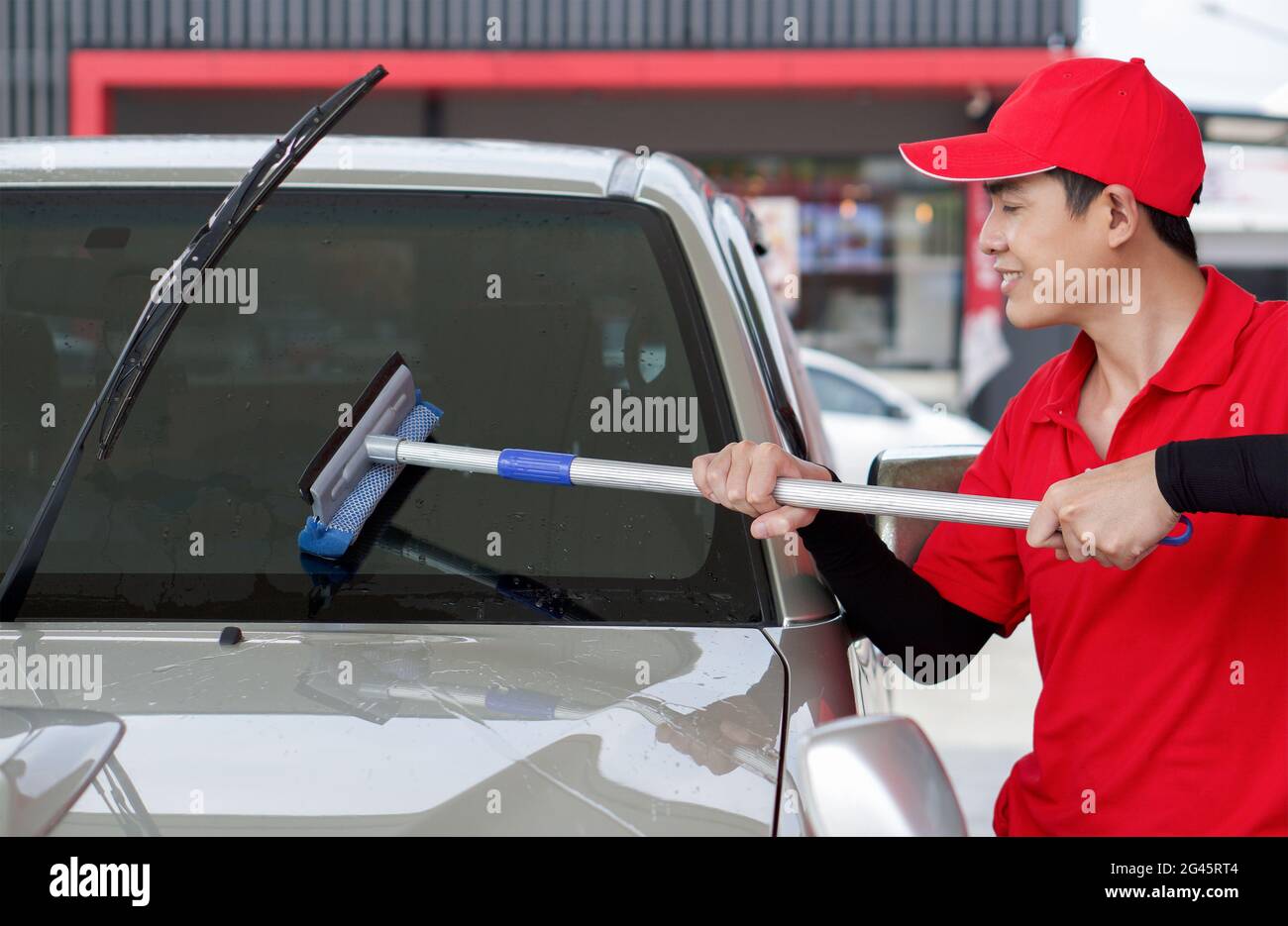 Ouvrier de station-service asiatique en uniforme rouge nettoyant le pare-brise de voiture avec raclette de fenêtre. Banque D'Images