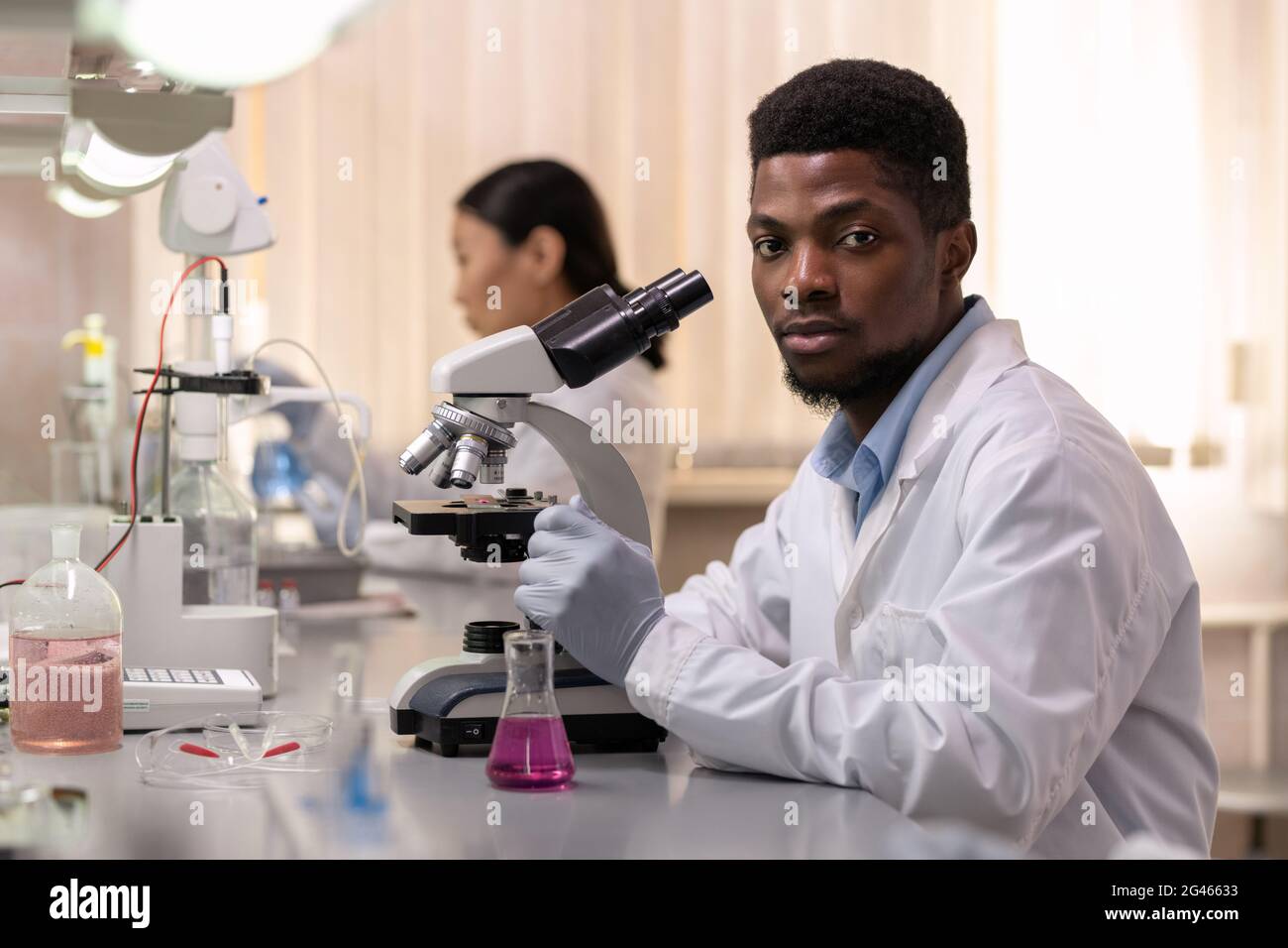 Jeune homme chimiste africain avec microscope assis au bureau en laboratoire Banque D'Images
