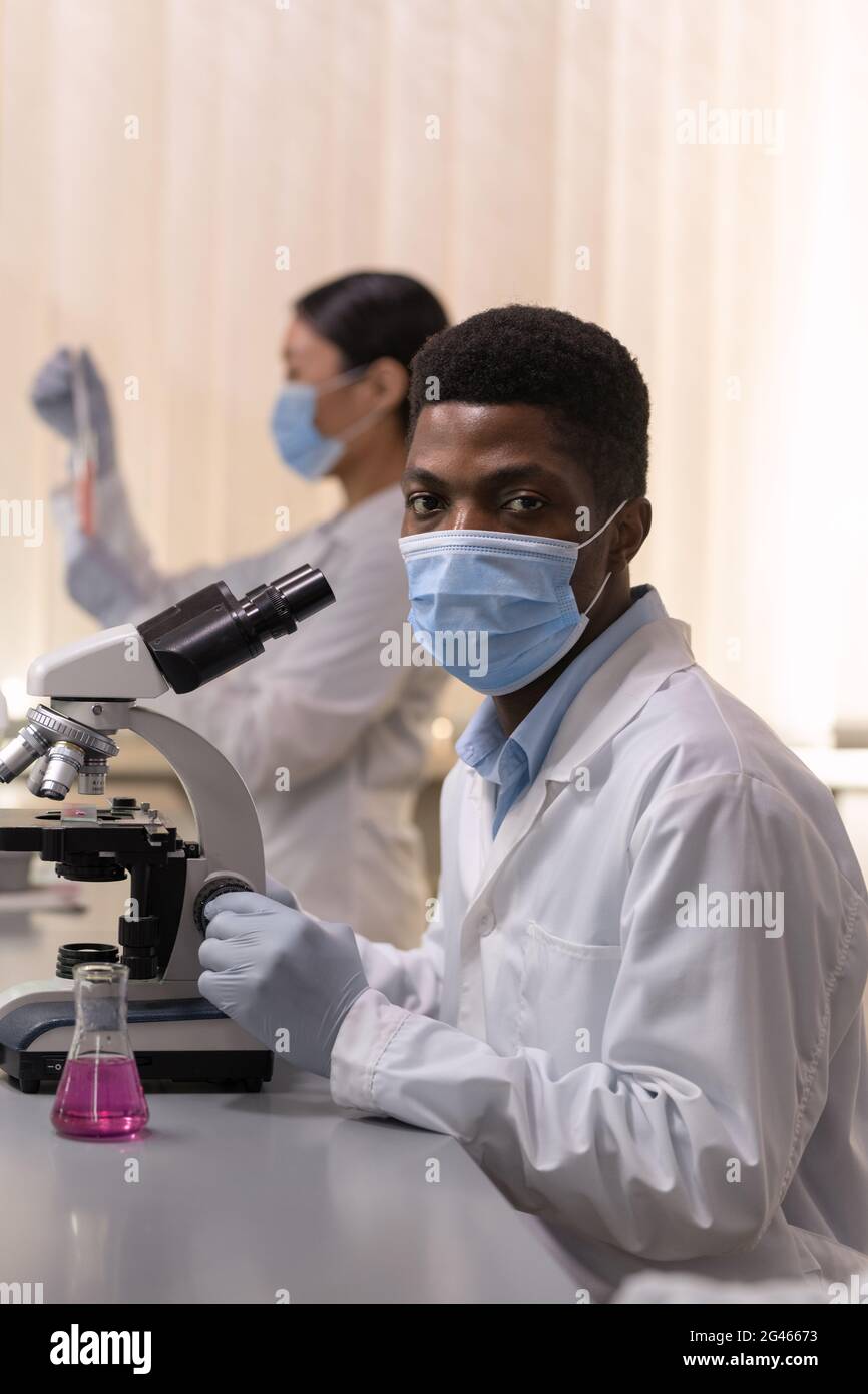 Chimiste masculin sérieux dans un masque de protection et un blouse de laboratoire travaillant avec un microscope Banque D'Images