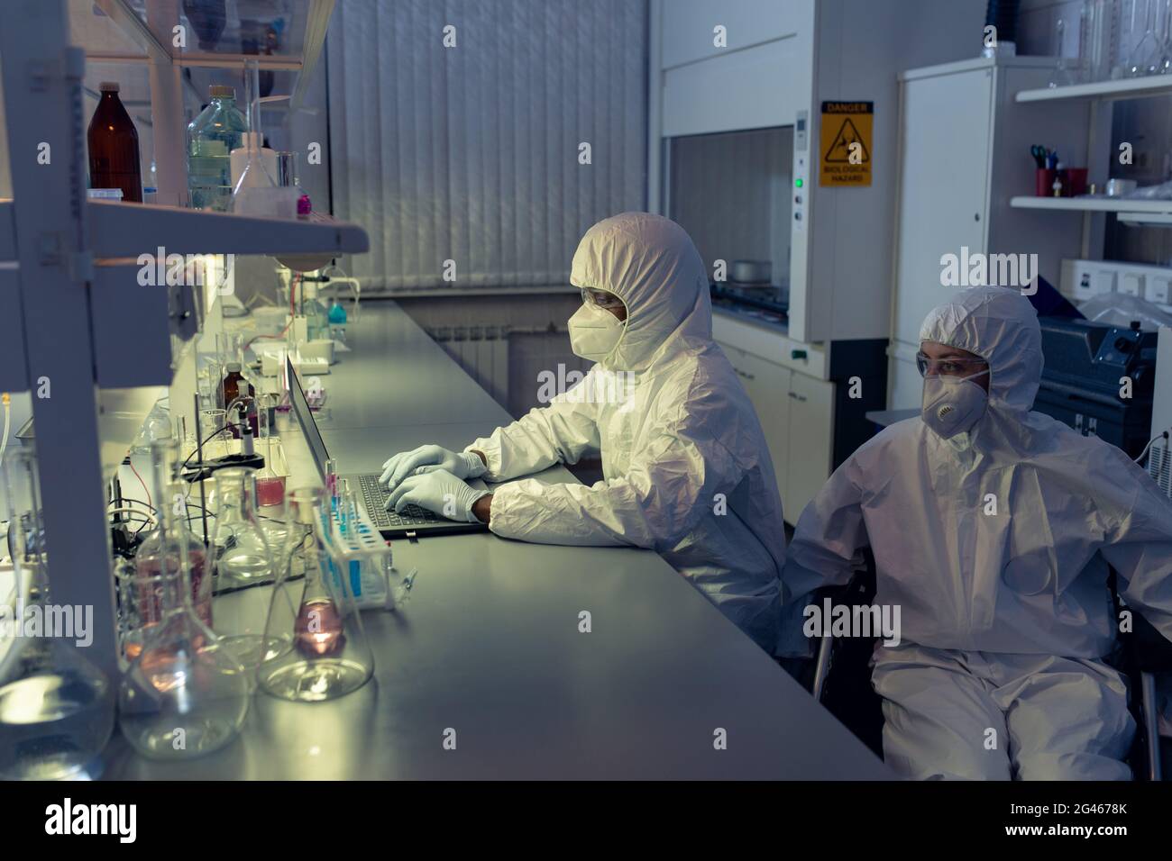 Femme scientifique en fauteuil roulant regardant l'écran de son ordinateur portable par sa collègue pendant son travail en laboratoire Banque D'Images