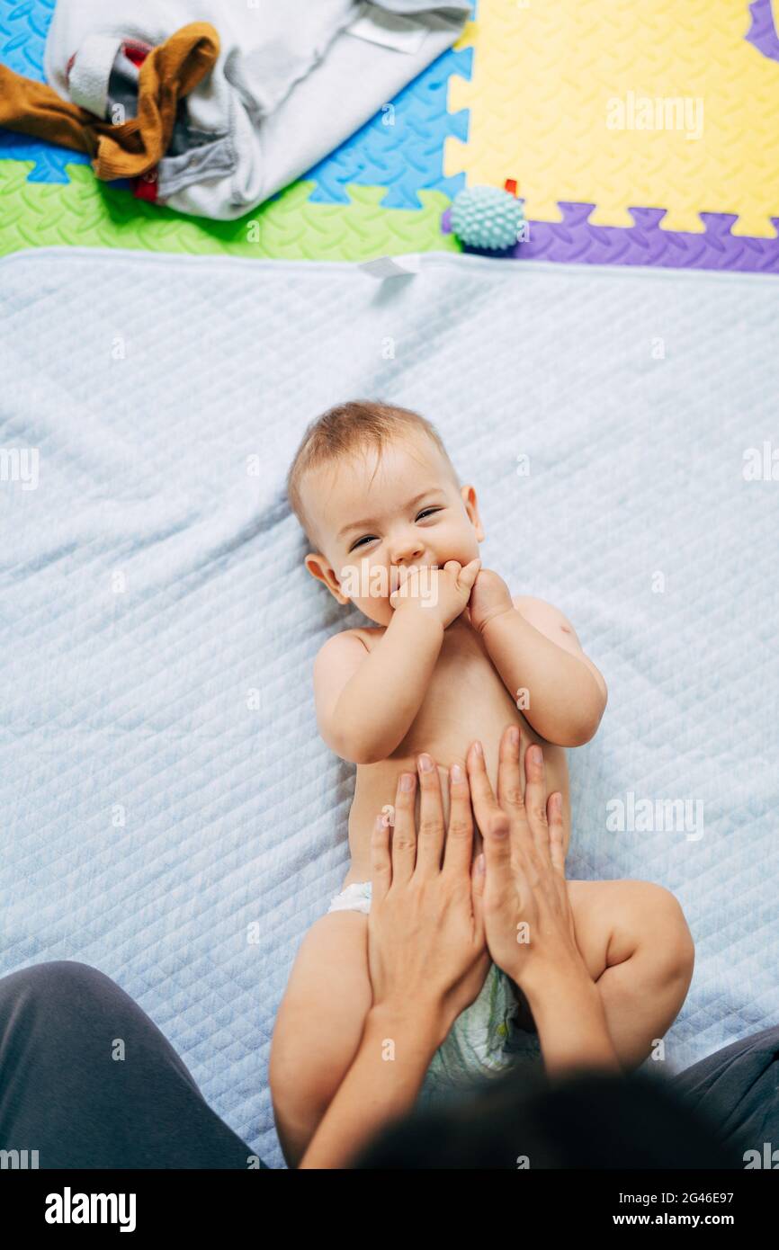 Bébé souriant dans une couche avec ses mains dans sa bouche repose avec son dos sur une couverture bleue sur un tapis coloré. Les mains de maman sont sur t Banque D'Images