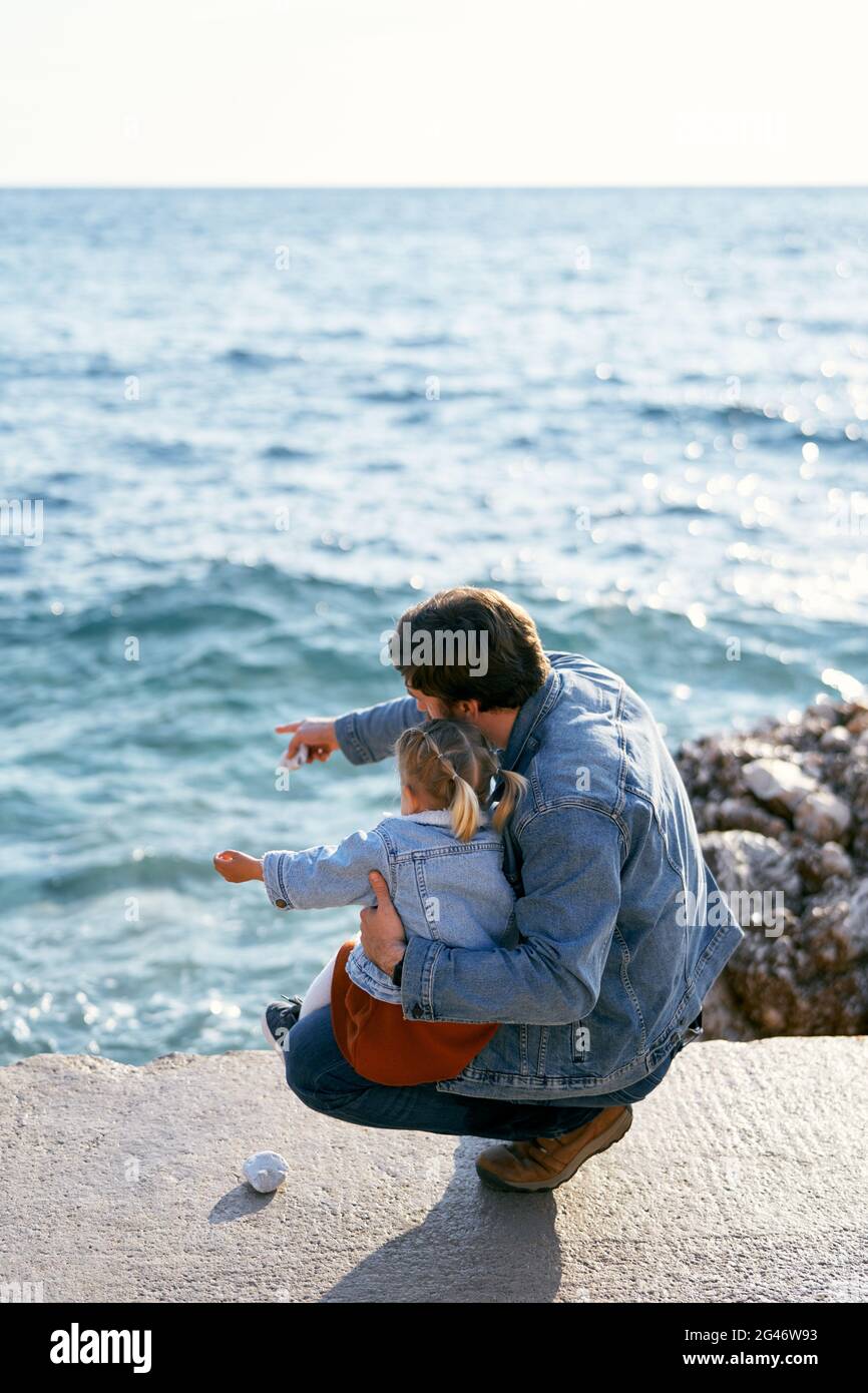 Papa s'est accroupi en tenant la petite fille sur ses genoux sur un grand rocher. Papa pointe son doigt sur la mer vers sa fille. Retour Banque D'Images