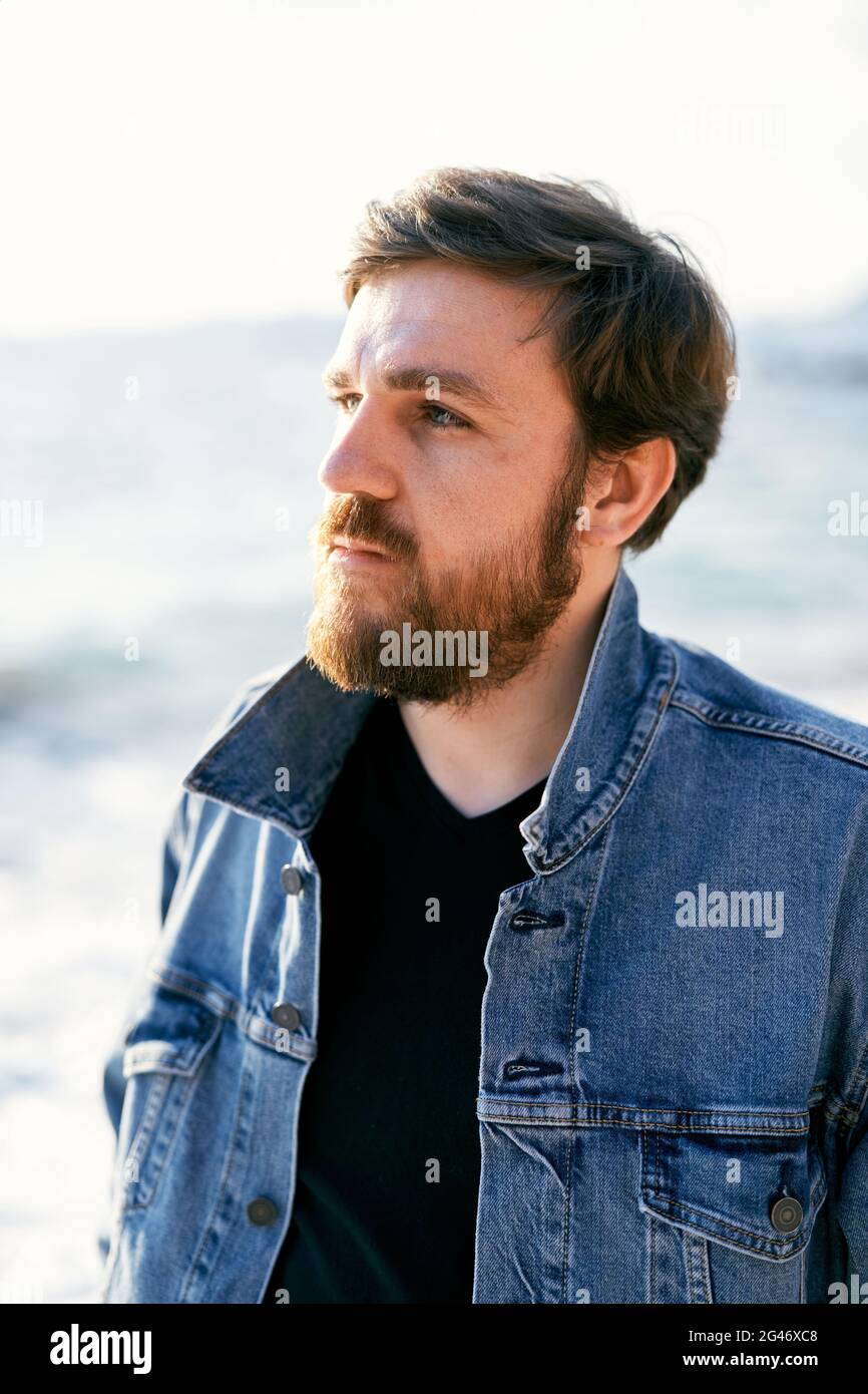 Portrait d'un homme pensif dans une veste en denim regardant la distance par rapport à l'arrière-plan de la mer. Gros plan Banque D'Images