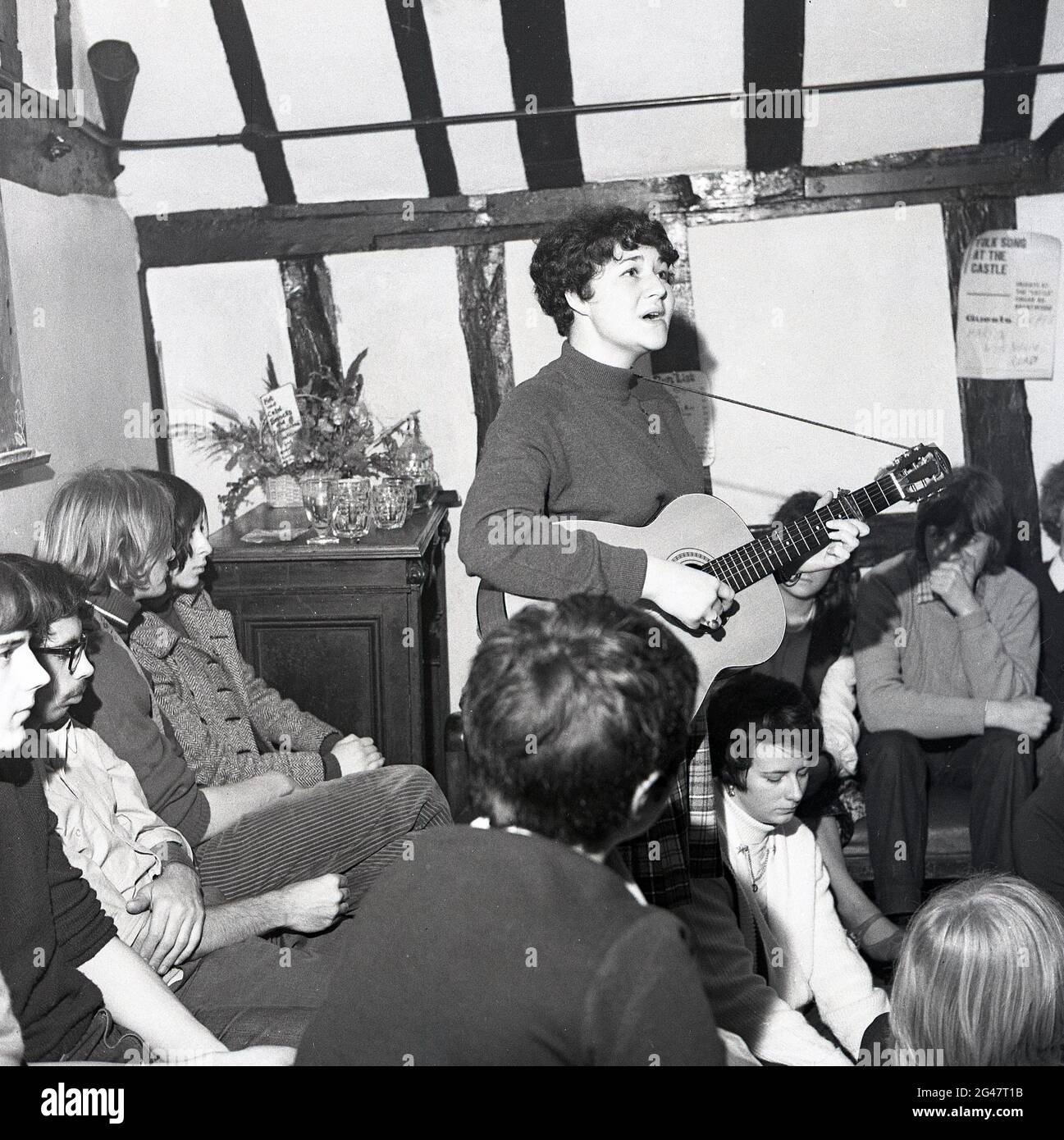 1970, musique folklorique historique, dans une salle d'un pub de campagne avec poutres en bois, un public écoutant une jeune chanteuse folklorique jouant une guitare et chantant une chanson au Castle Inn, Colchester, Essex, Angleterre, Royaume-Uni. Banque D'Images