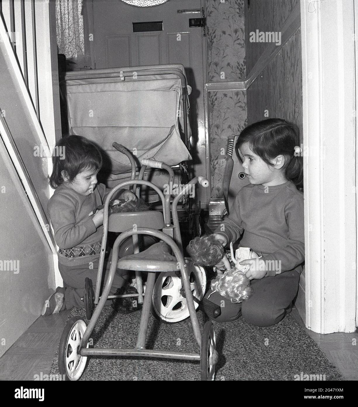 Années 1960, historique, à l'intérieur du couloir d'une maison mitoyenne, surpeuplée d'un landau et de tricycles métalliques pour enfants, deux petits enfants, pressés à côté des escaliers mur d'abd, agenouillé sur le sol, jouant avec des jouets et des poupées doux, Angleterre, Royaume-Uni. Banque D'Images
